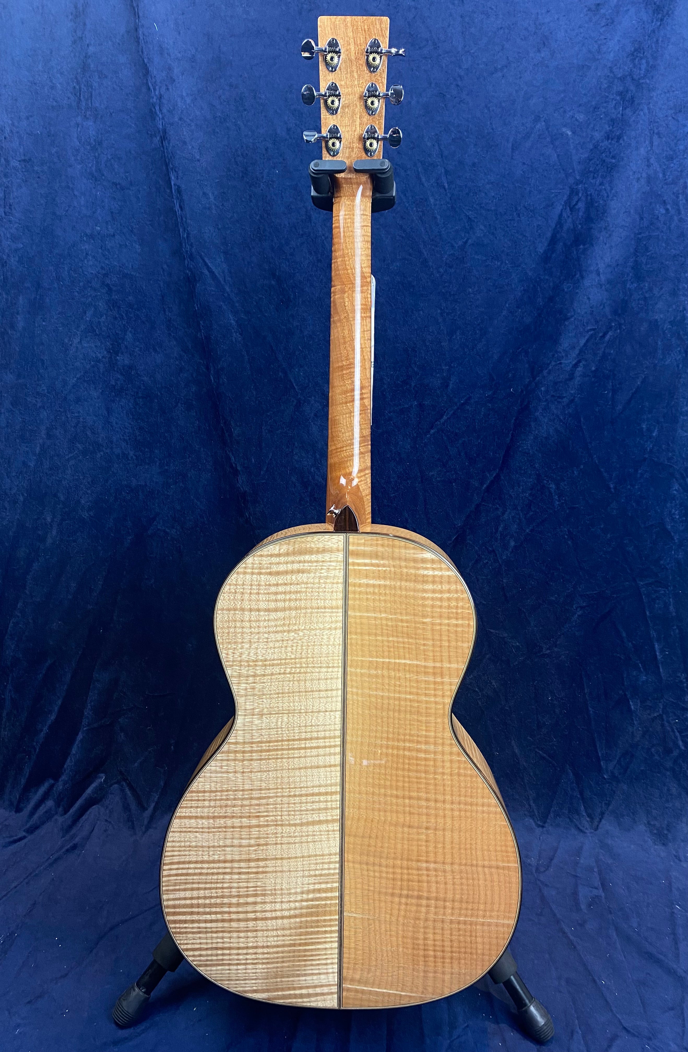 Back view of an acoustic guitar with a glossy, striped light wood finish, six tuning pegs, and a central seam running vertically down the body.