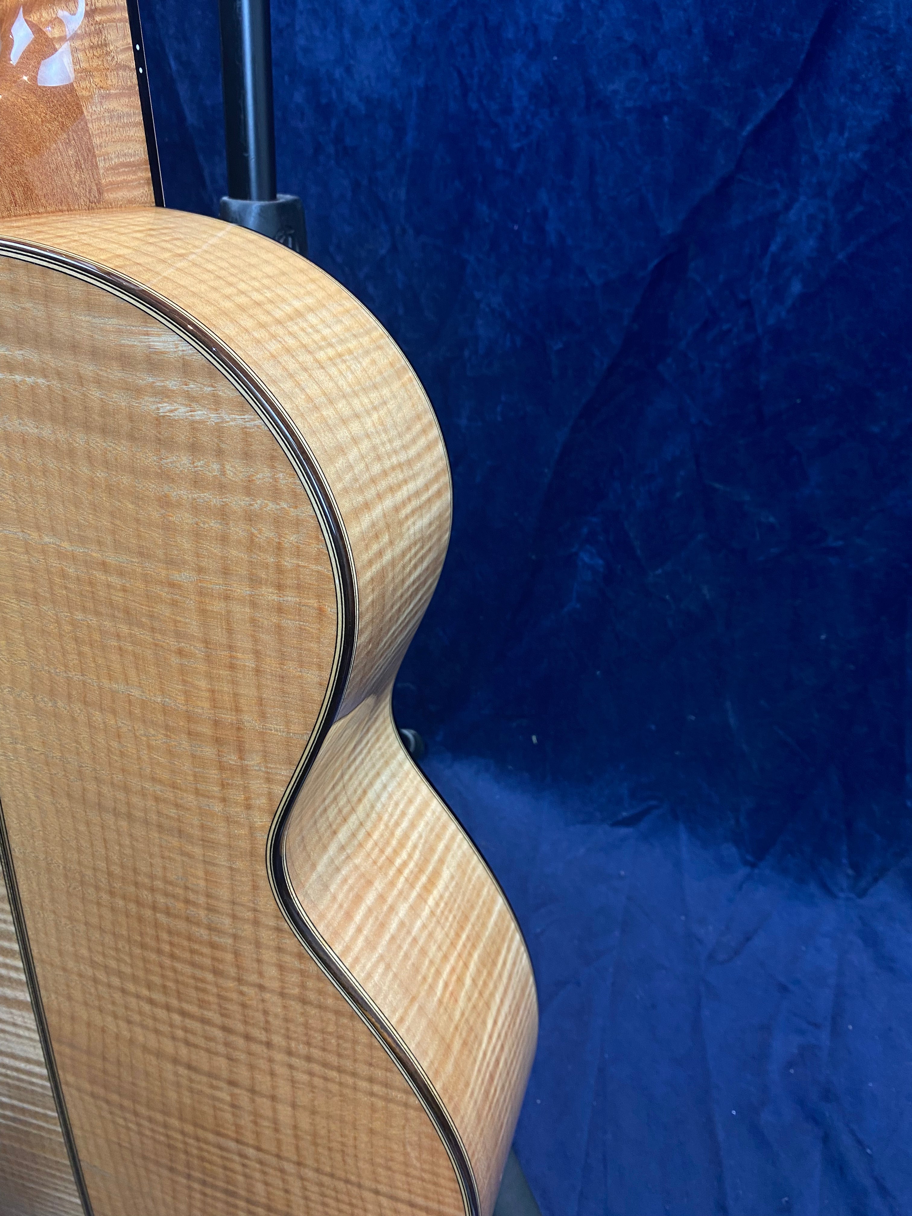 Curvy side and back of an acoustic guitar showing light-colored, striped wood grain against a dark blue background.