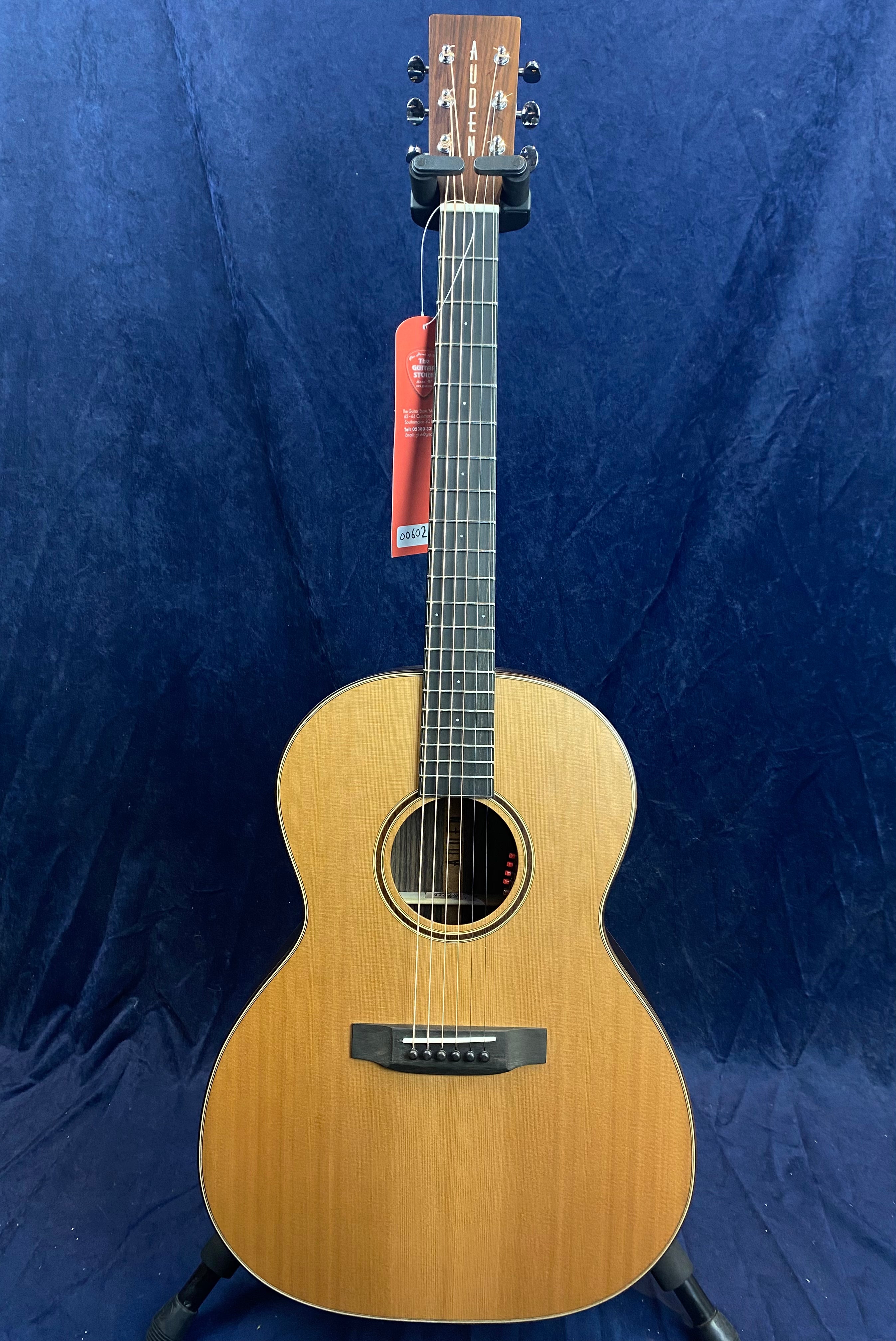Auden brand acoustic guitar with a natural wood finish, six strings, and a rounded body, displayed upright on a stand against a dark blue background.