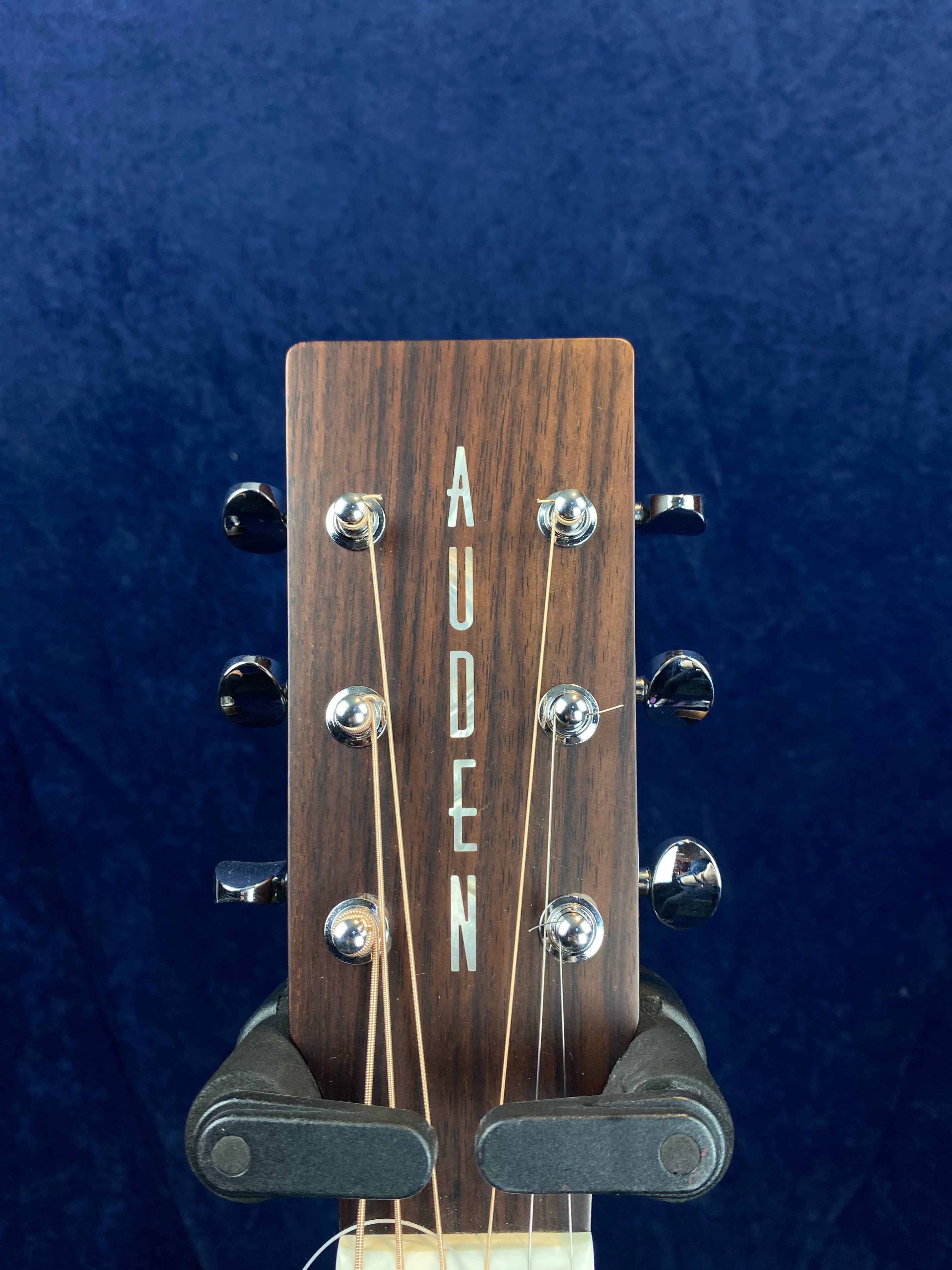 Headstock of an acoustic guitar with six silver tuning pegs and the word AUDEN inlaid vertically on dark wood.