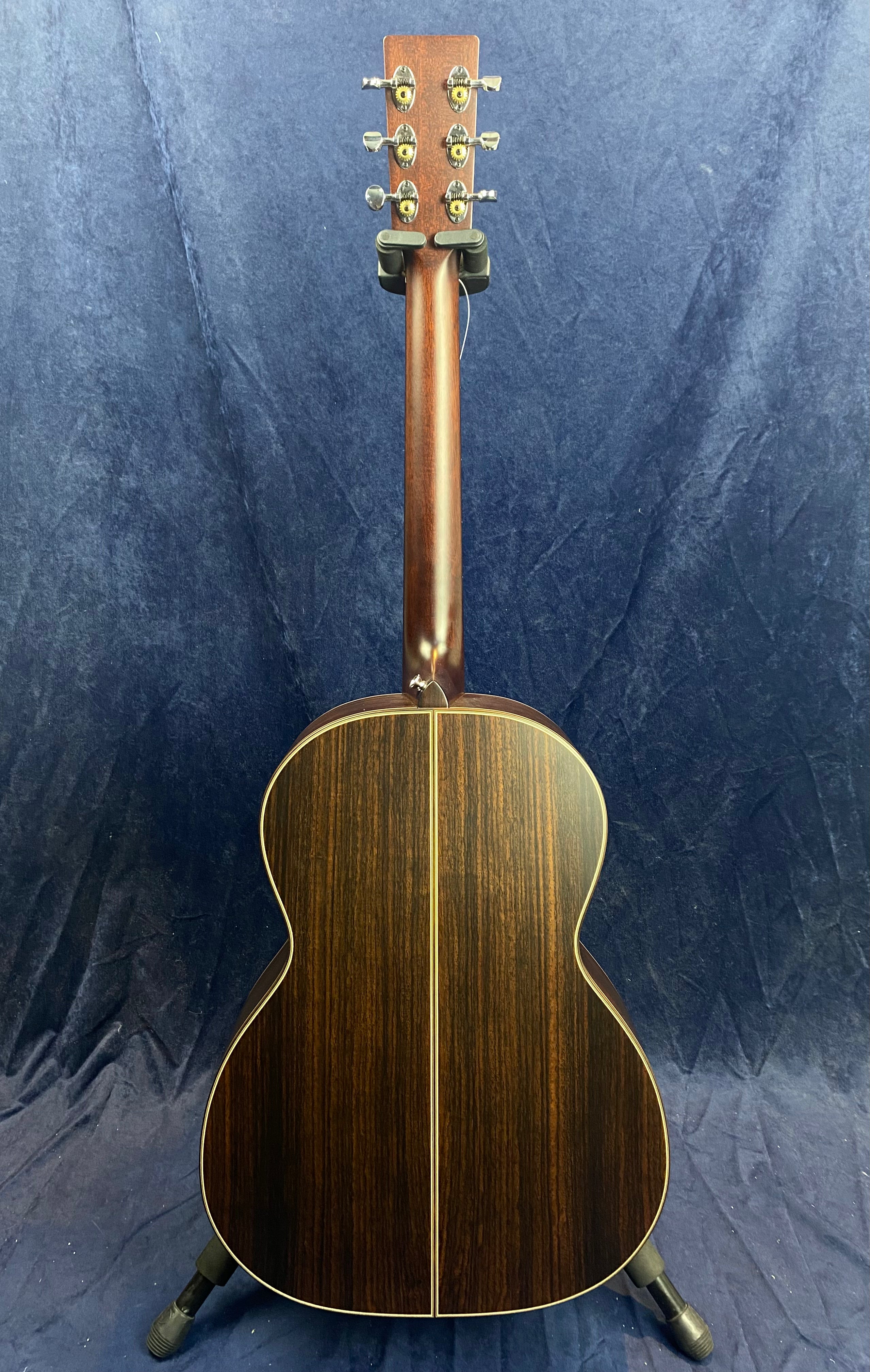 The back of an acoustic guitar with dark wood, light center stripe, and silver tuning pegs, standing upright against a blue cloth background.