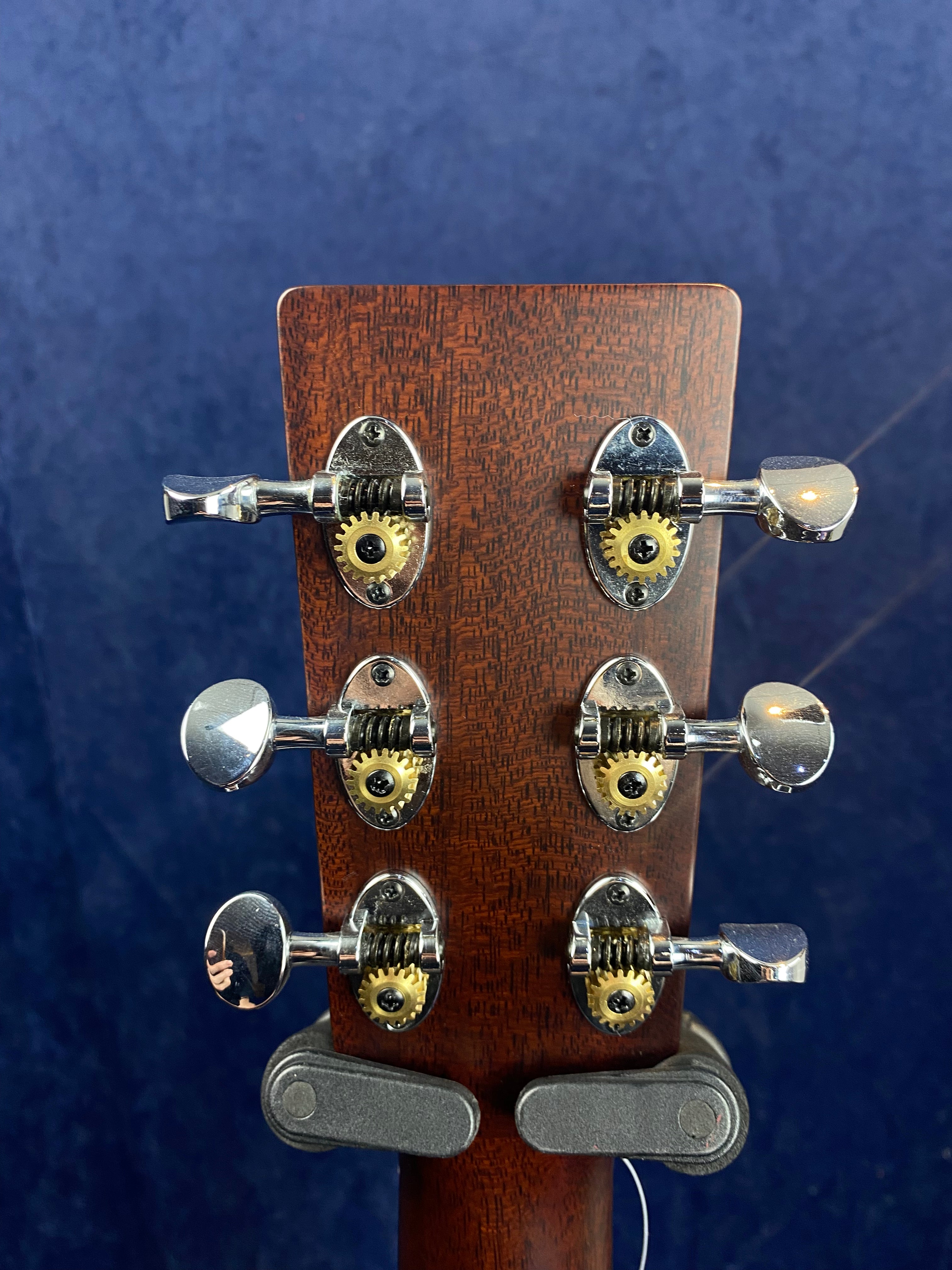 Rear headstock of an acoustic guitar showing three pairs of chrome and brass tuning pegs against a dark blue background.