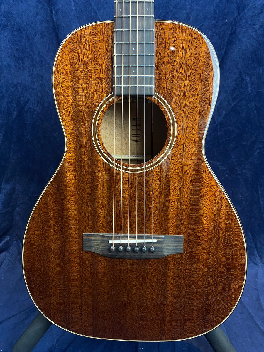 Small-bodied acoustic guitar with a rich, dark wood grain finish, six steel strings, a round soundhole, and a black bridge with white pin inlays.