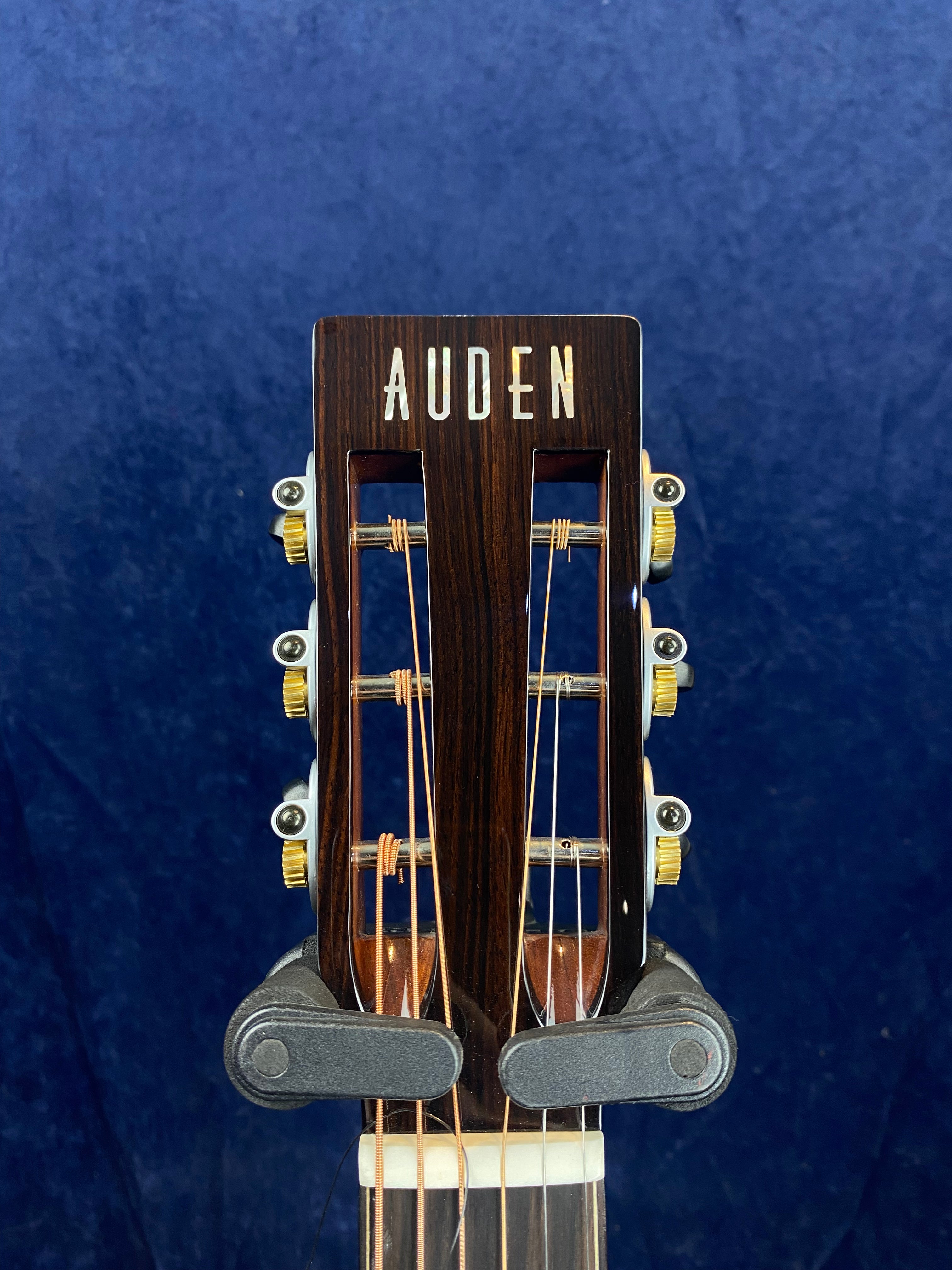 Slotted headstock of an Auden acoustic guitar with gold tuning pegs, viewed against a dark blue background.