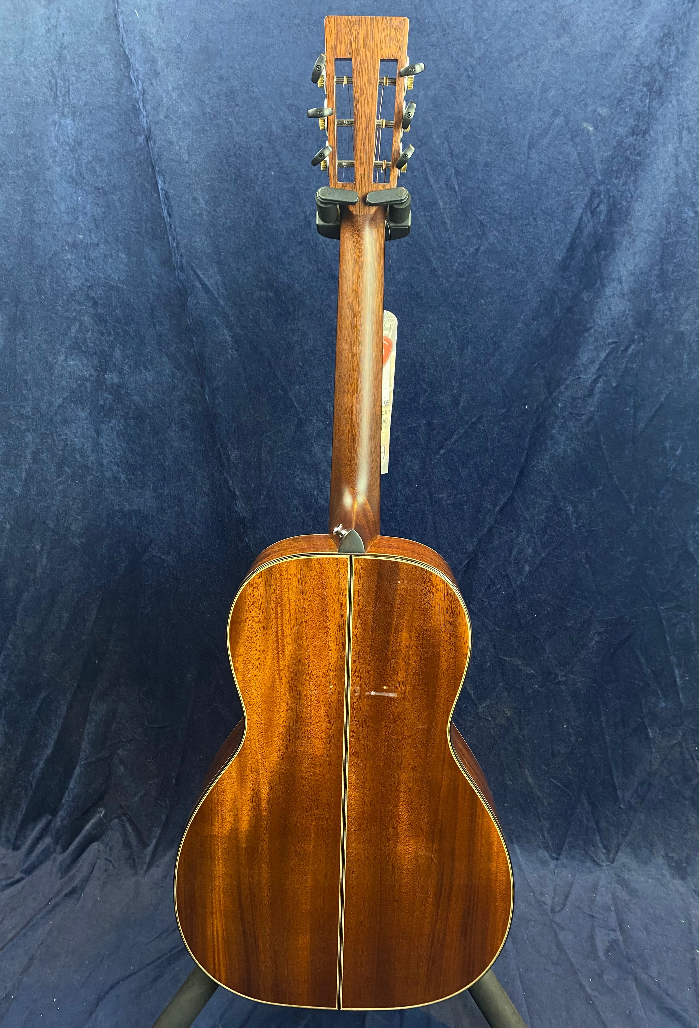 Mahogany acoustic guitar shown from the back, with a central seam and a glossy finish, supported upright on a stand against a dark blue background.