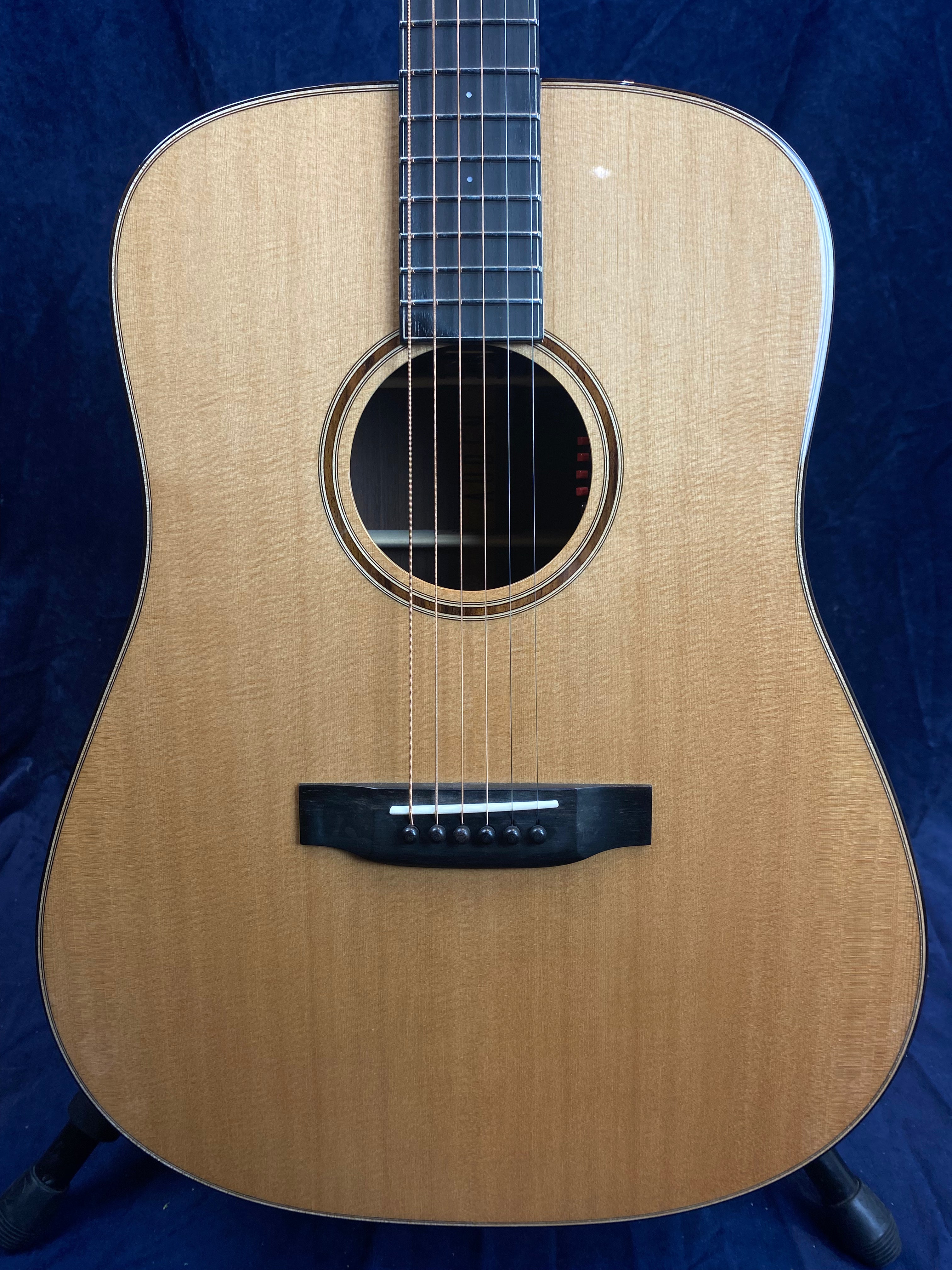 Natural wood-finish acoustic guitar with six strings, black bridge, round sound hole, and a dark fingerboard set against a dark blue background.