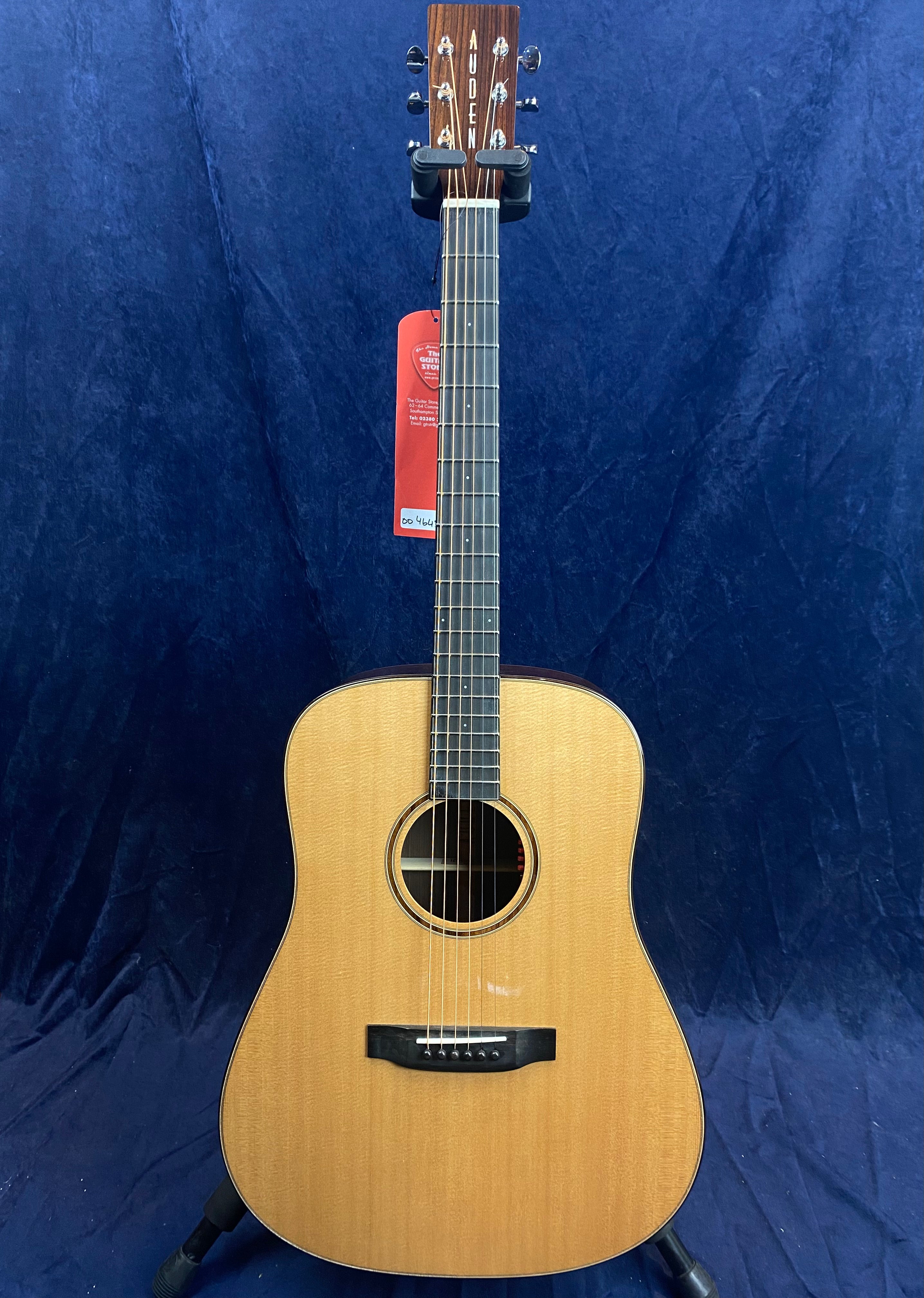 Auden acoustic guitar with a natural wood finish, dark fretboard, and six strings, displayed on a stand against a dark blue background.