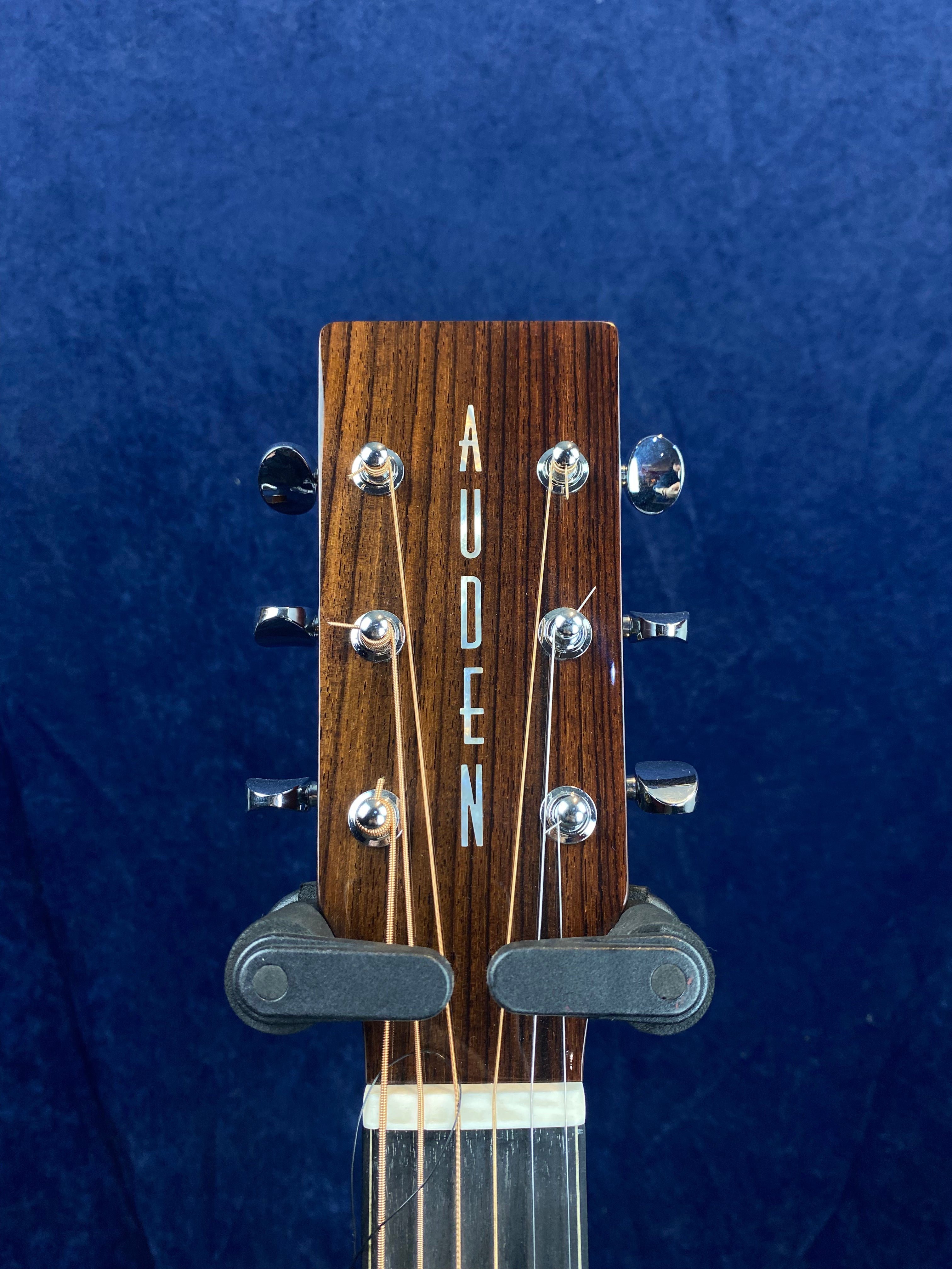 Close-up of an Auden acoustic guitar headstock with six tuning pegs, vertical logo lettering, and strings visible against a dark blue background.