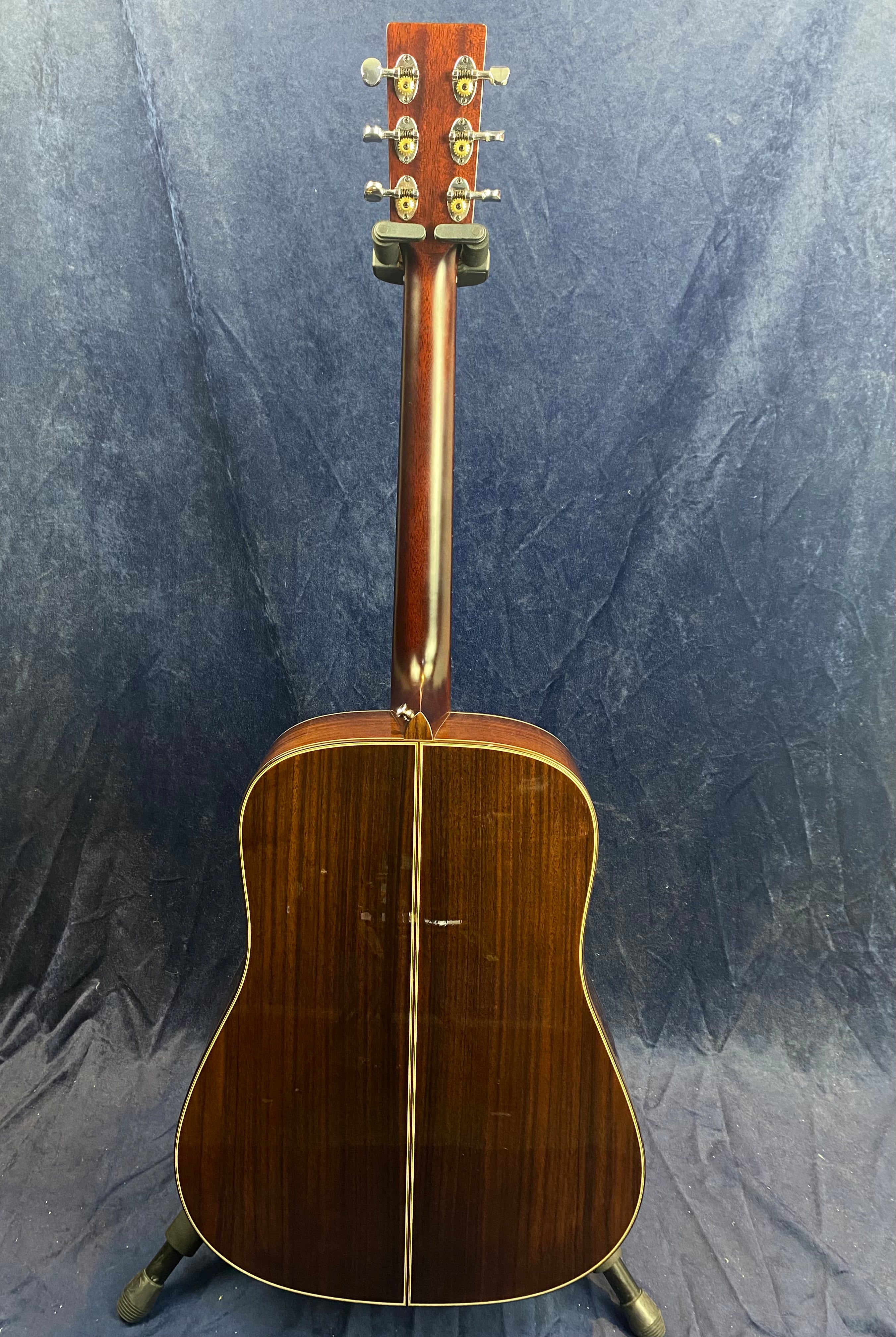 Back view of an acoustic guitar with a glossy dark wood finish, visible grain, gold tuning pegs, and a faint scratch near the lower center.