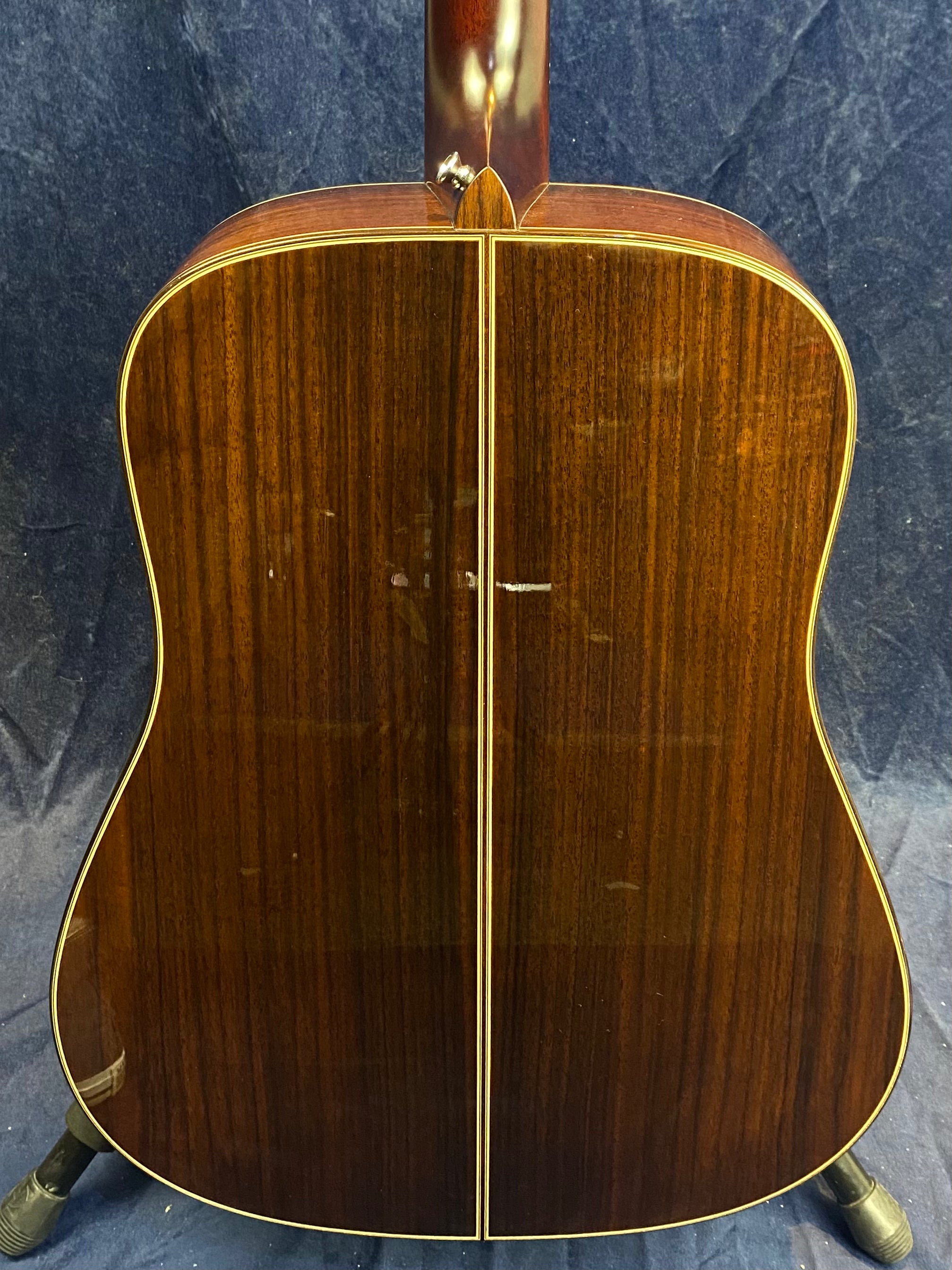 The back of an acoustic guitar made of dark, polished wood with a light center seam and thin yellow edge binding, shown upright on a stand.