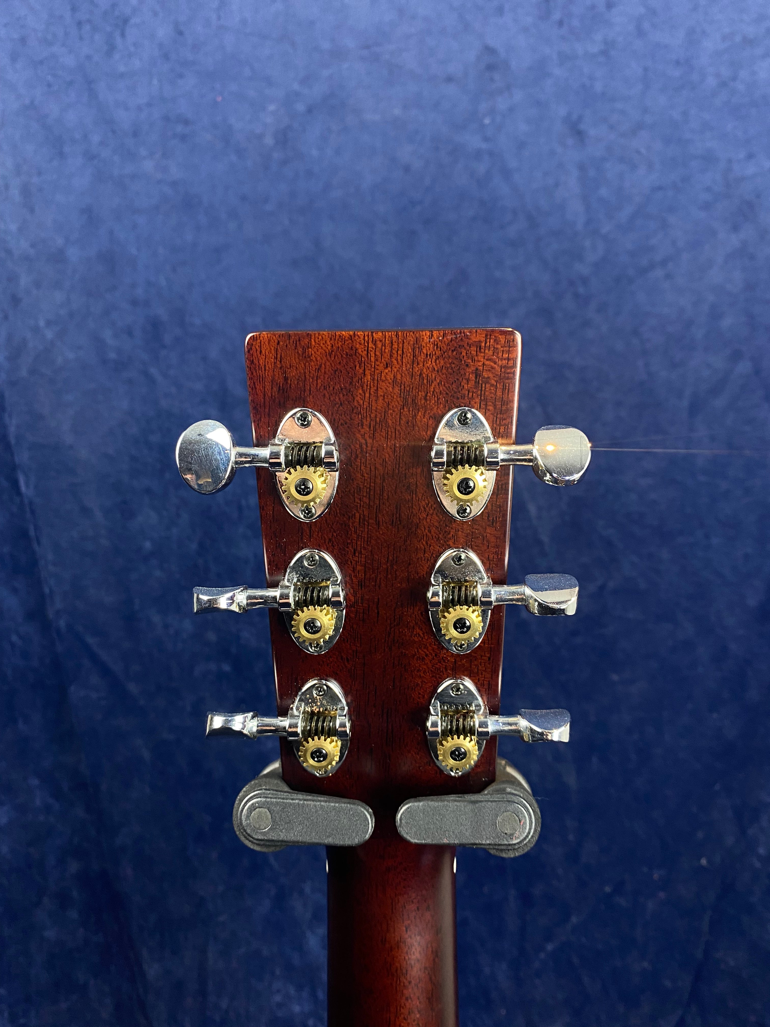Back of an acoustic guitar headstock showing six chrome tuning pegs with exposed gears against a dark blue background.