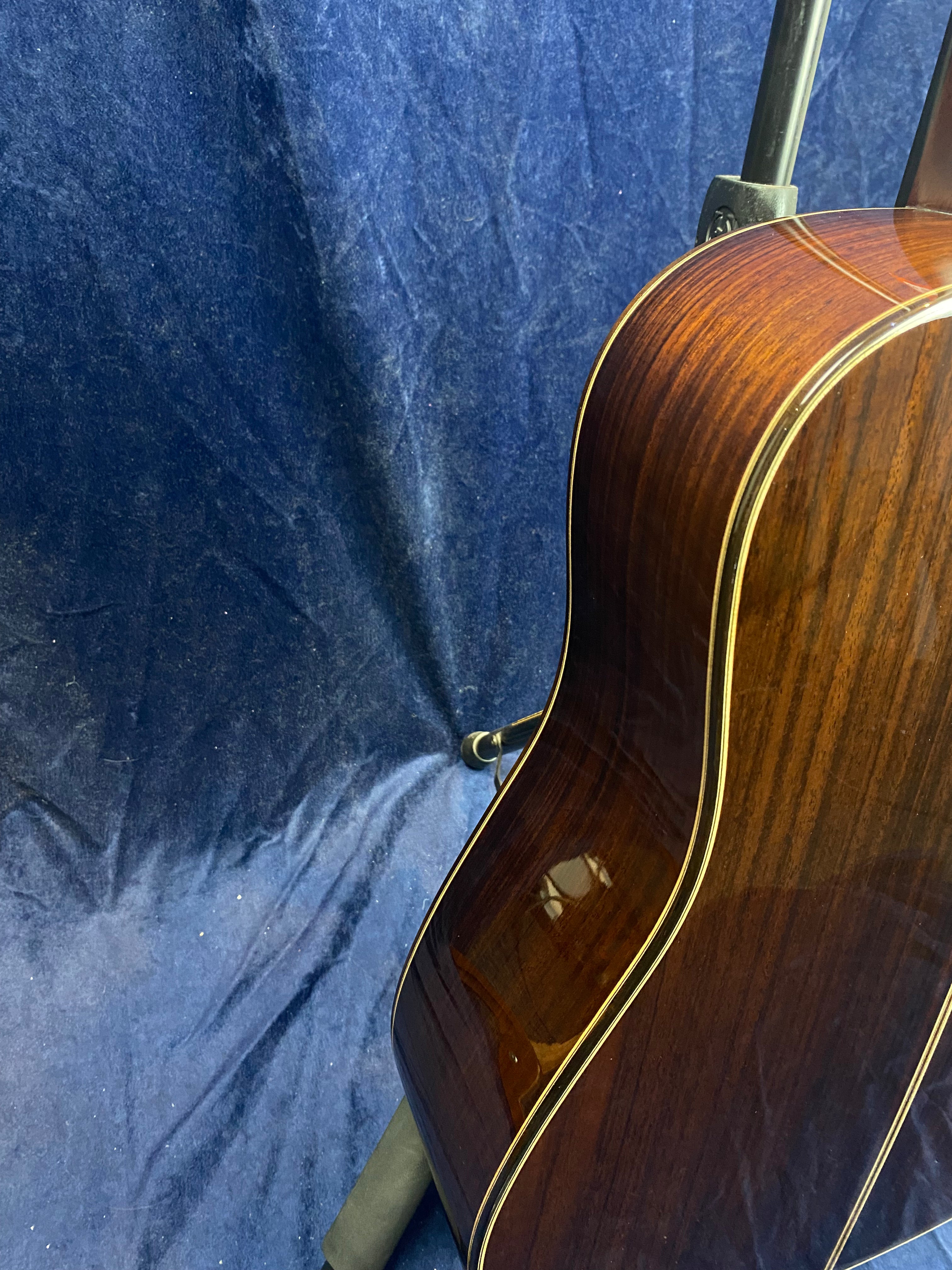 Glossy dark wood acoustic guitar body with light binding, shown from the side against a blue fabric background, guitar is on a black stand.