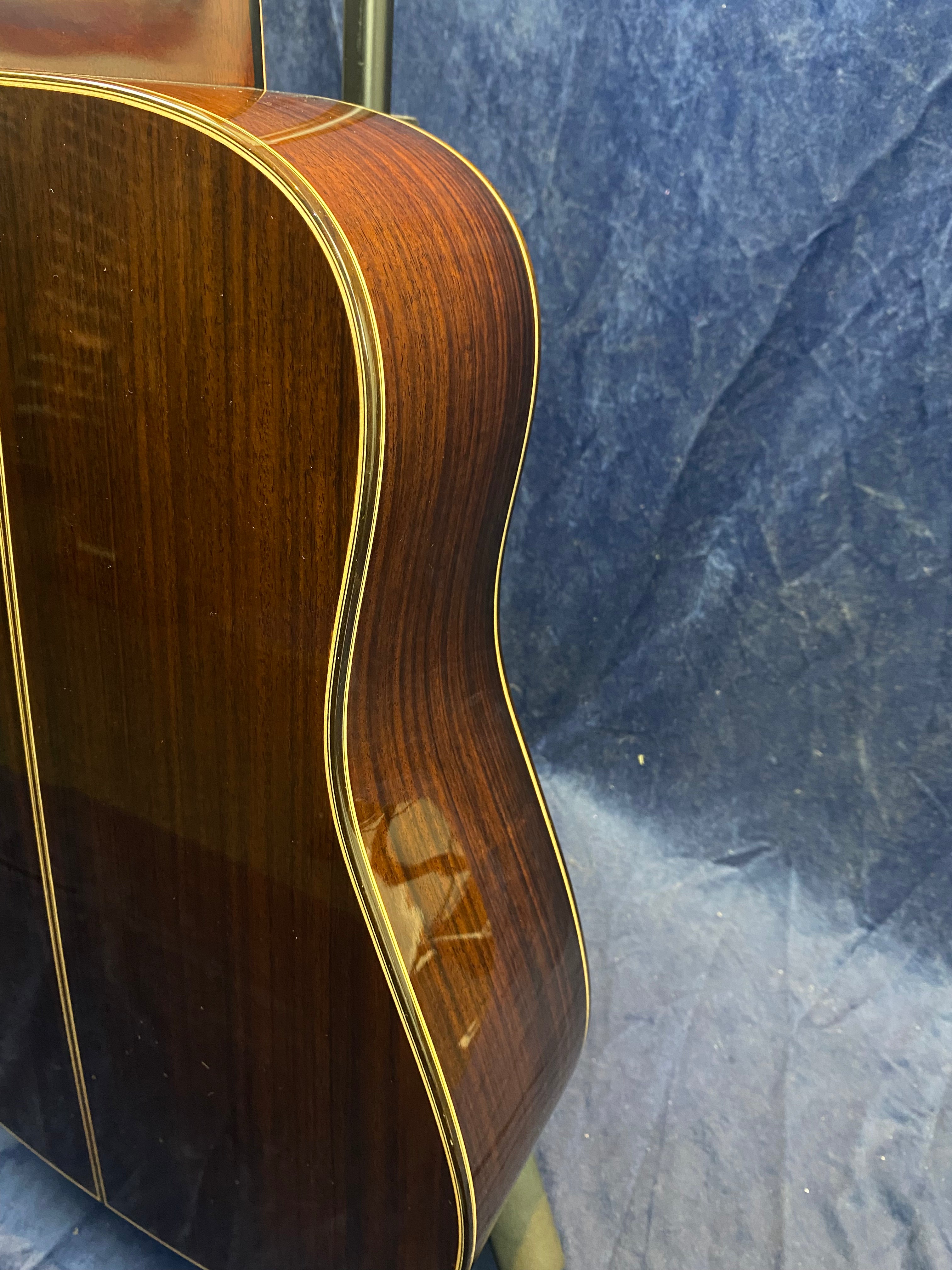 Glossy dark wood acoustic guitar body with light-colored binding, shown from the side against a textured blue background.