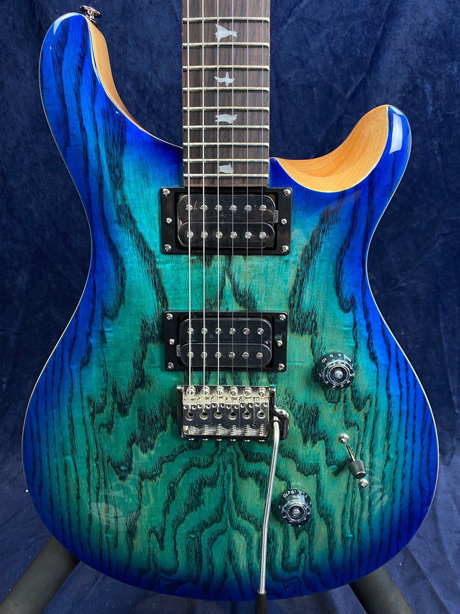 Electric guitar with a blue-green burst wood grain finish, two humbucker pickups, three control knobs, a switch, and a tremolo bridge.