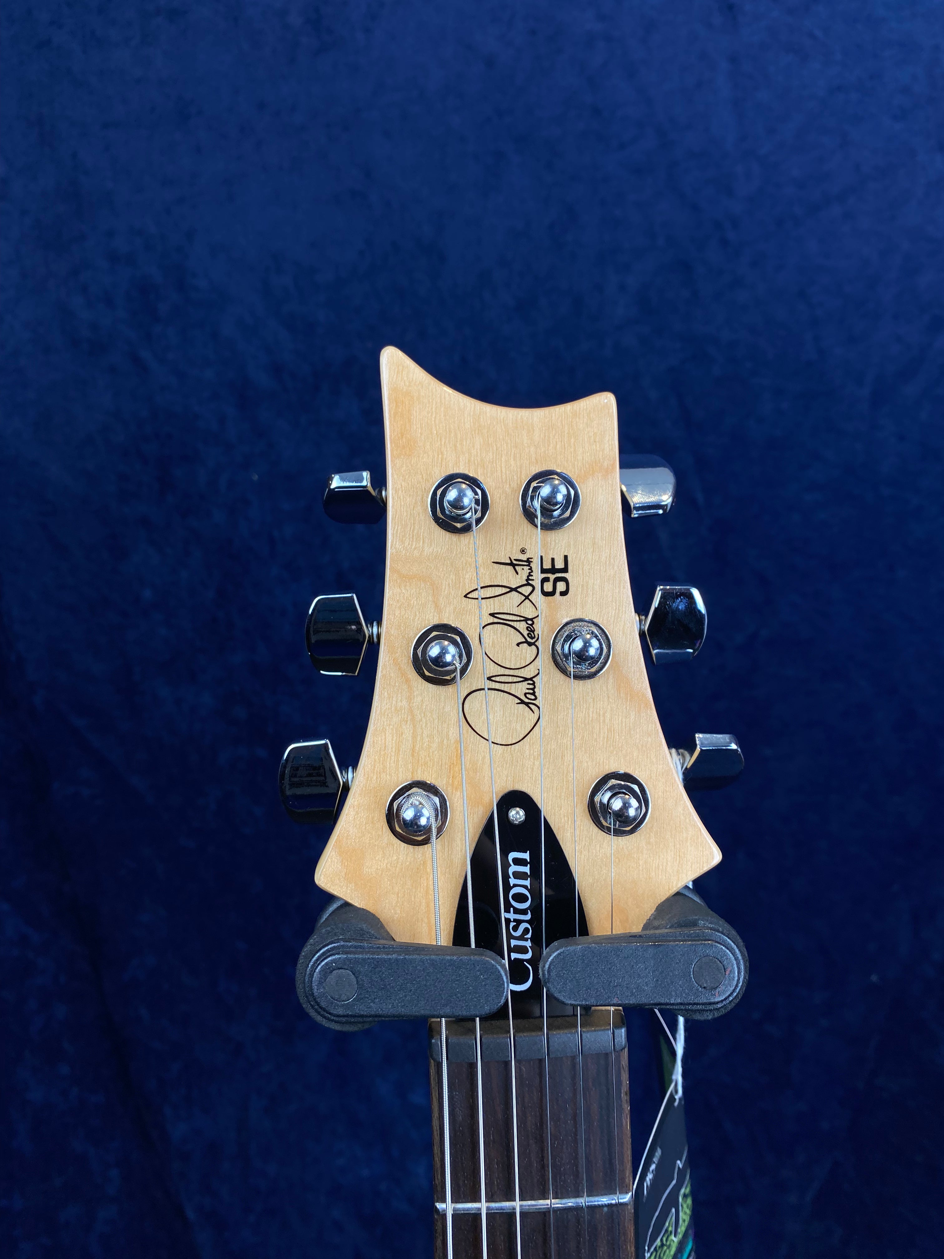Electric guitar headstock with six tuning pegs, natural wood finish, PRS SE series logo, and "Custom" truss rod cover on dark blue background.