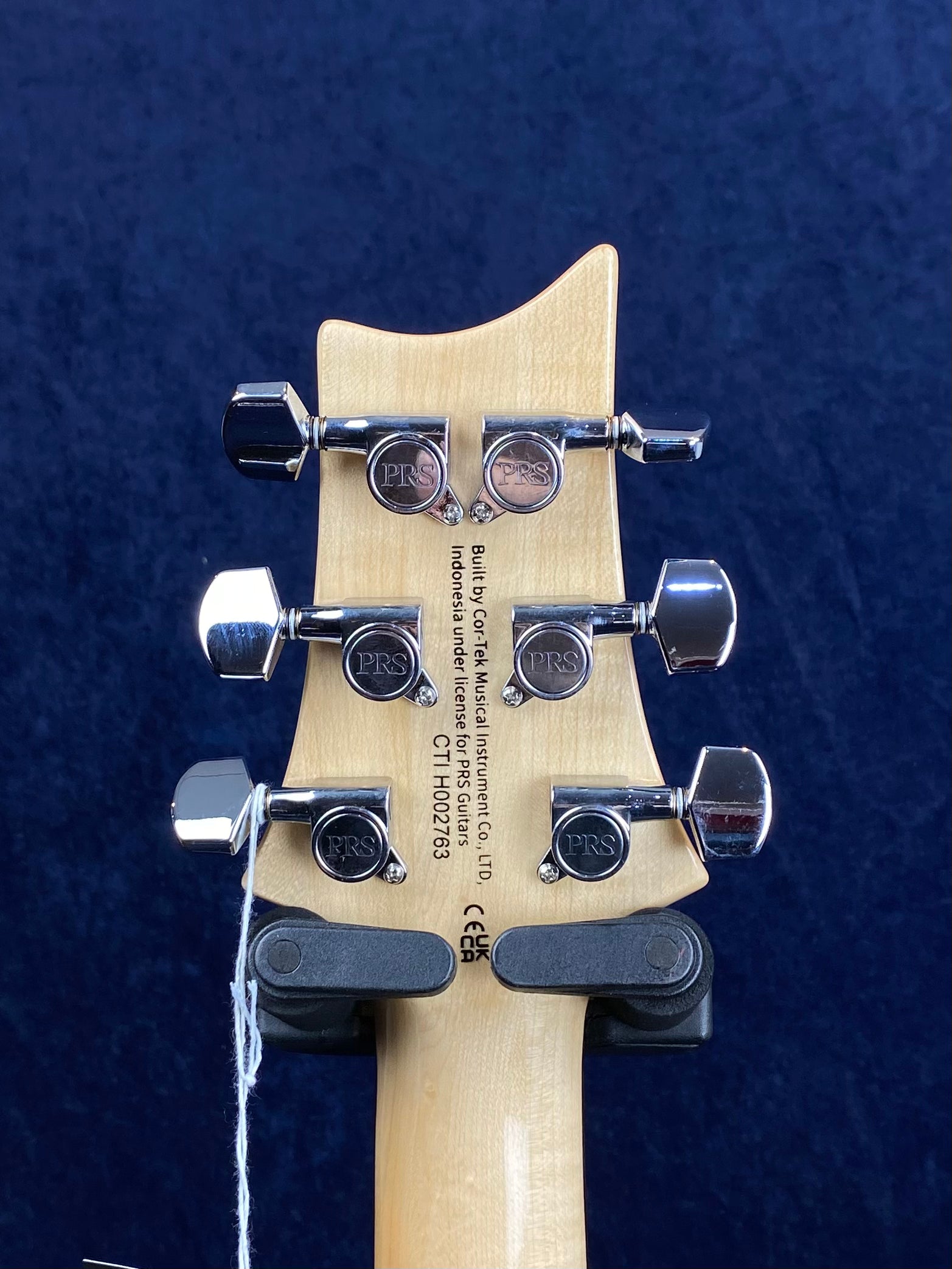 Back of a PRS electric guitar headstock showing six chrome tuning pegs, PRS logos, and manufacturer info printed on the light wood.