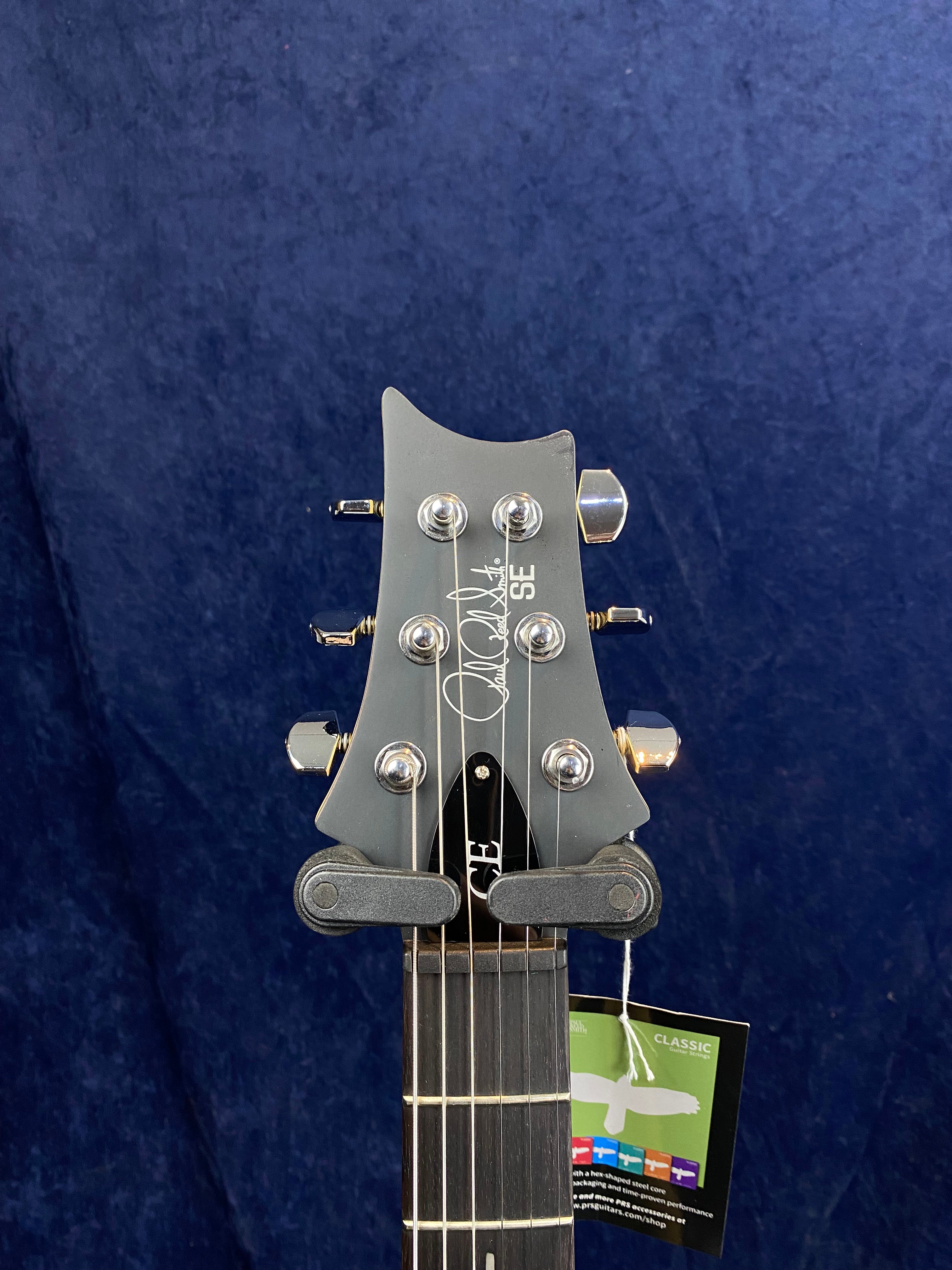 The headstock of a PRS SE electric guitar with six tuning pegs, silver hardware, and a tag attached below the nut.