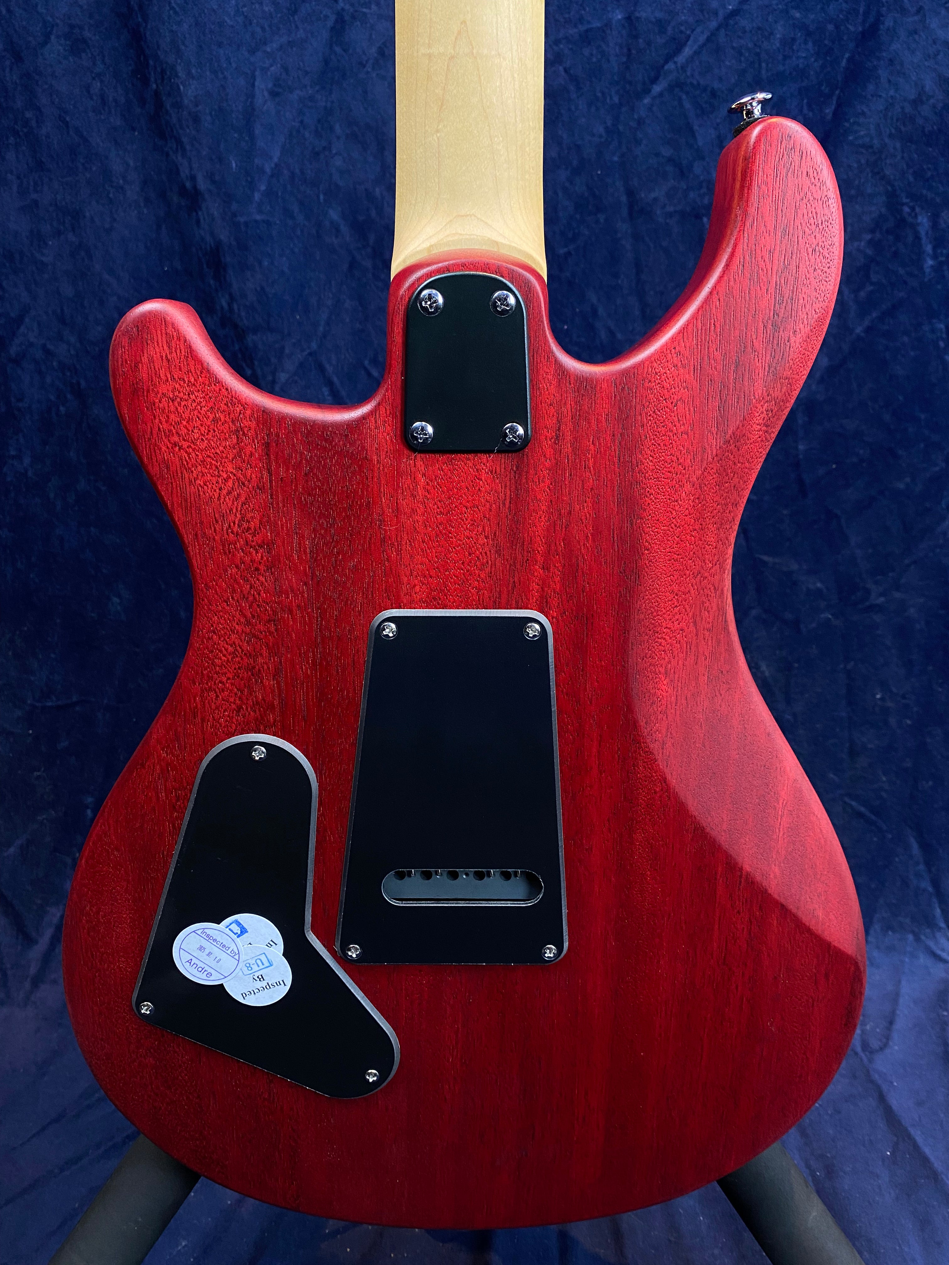 Back of a red electric guitar with a natural wood neck, black plastic cover plates, and inspection stickers on the lower cover.