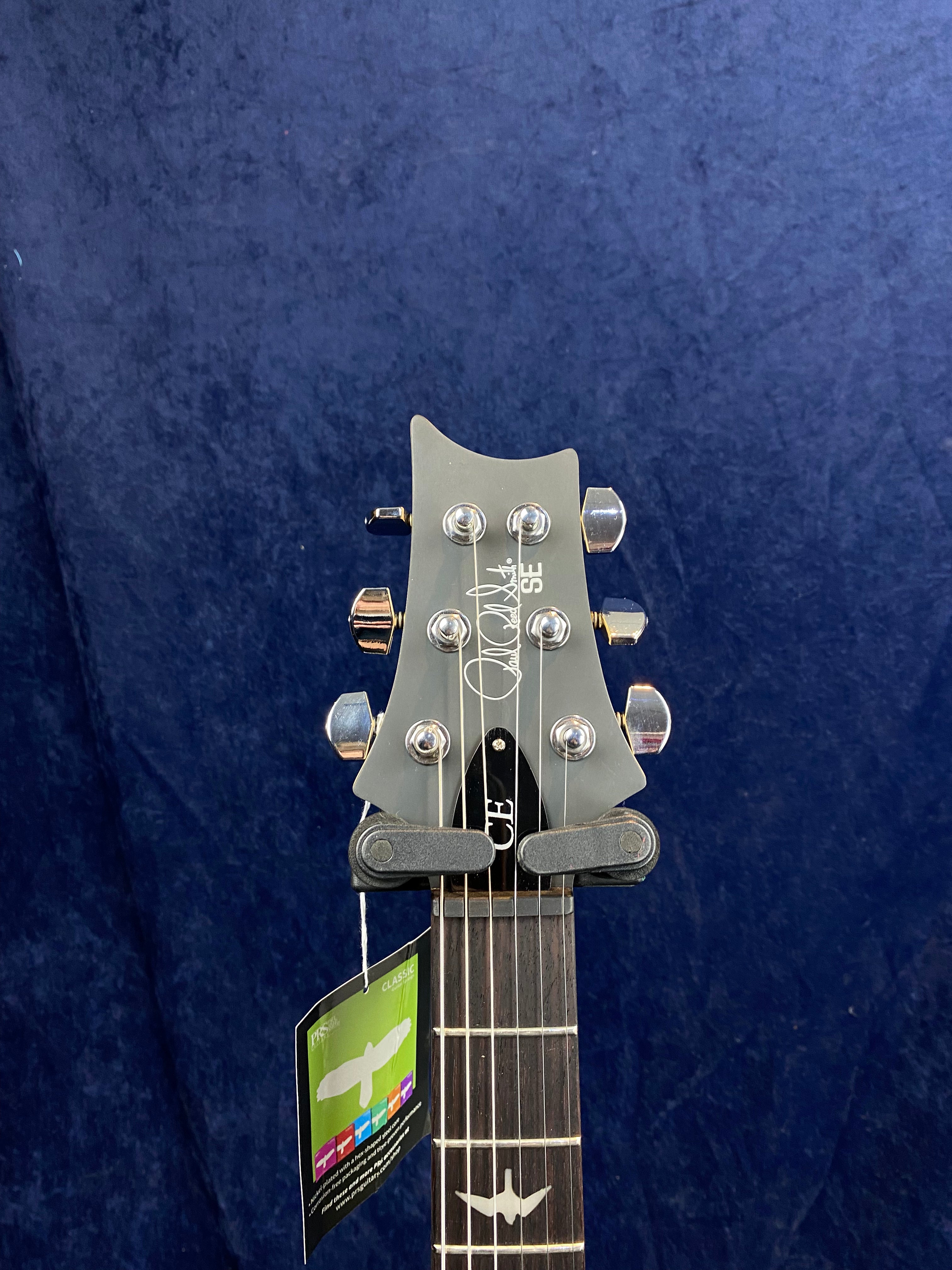 Electric guitar headstock with six tuning pegs, PRS SE logo, and a hang tag; dark wood fretboard with bird inlays visible.