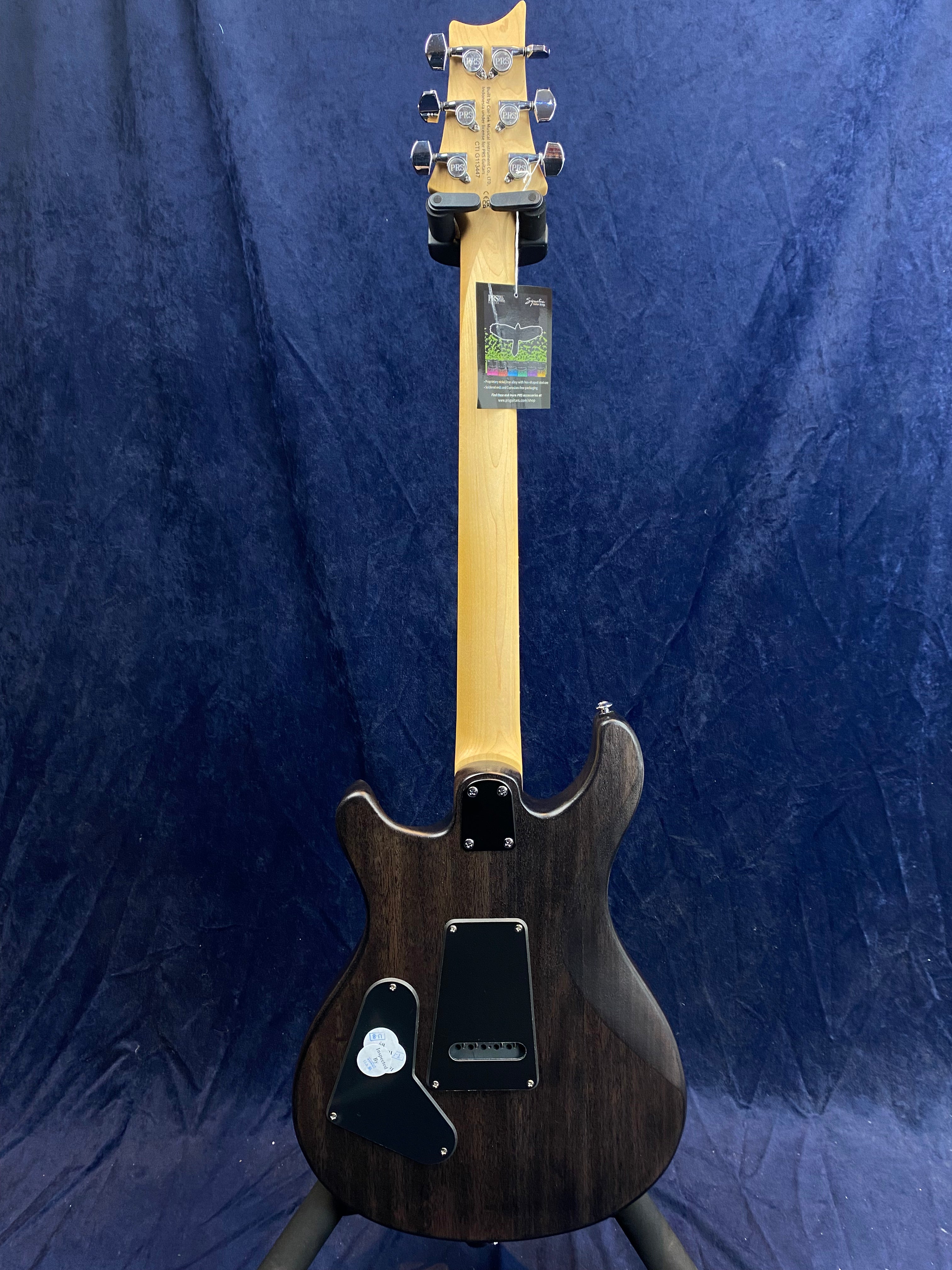 Electric guitar shown from the back with a natural wood neck, dark body, chrome tuners, and black back plates. A tag hangs near the headstock.