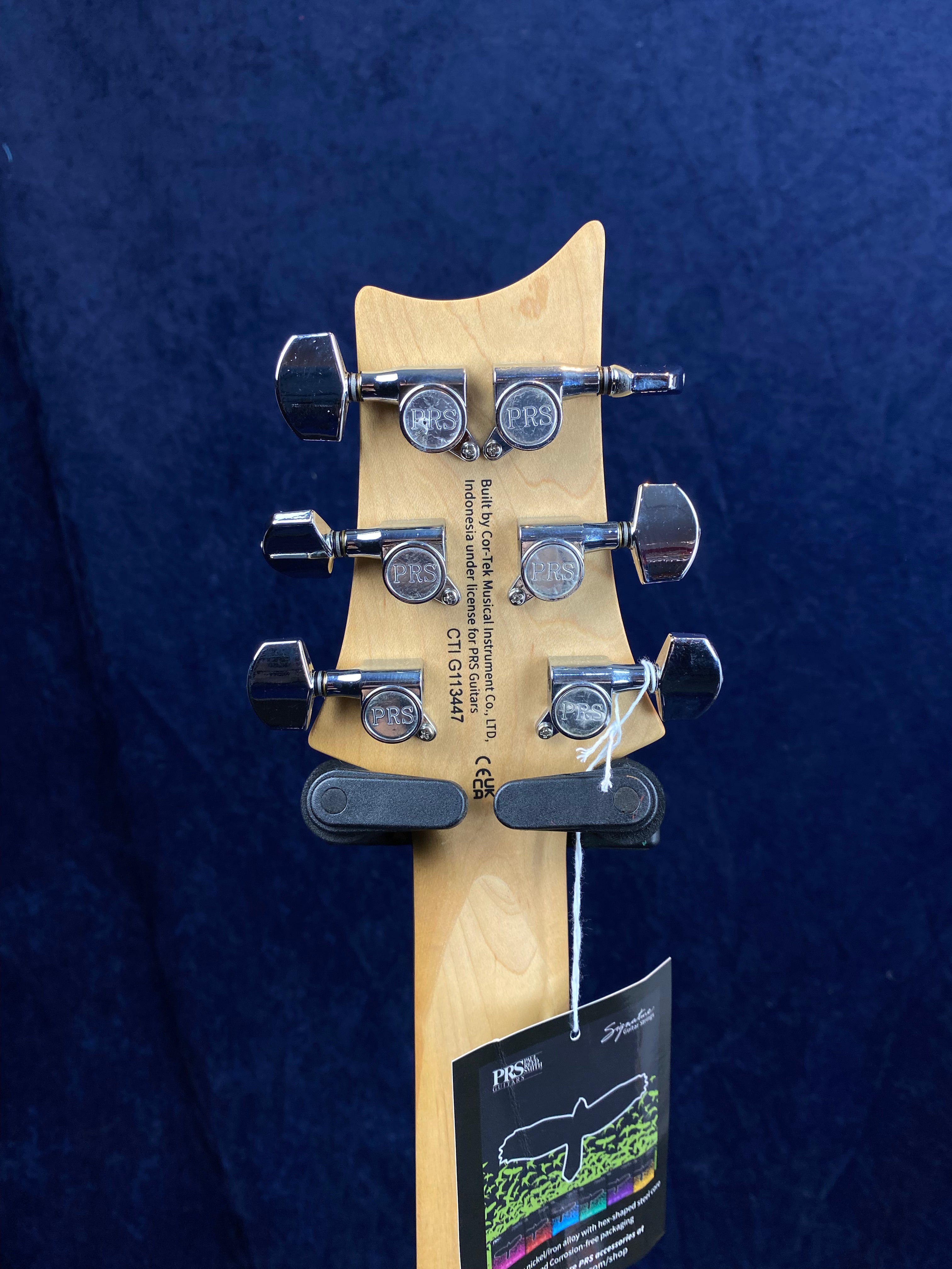 Back of a PRS electric guitar headstock showing six chrome tuning pegs, manufacturer info, a string tag, and a natural wood finish.