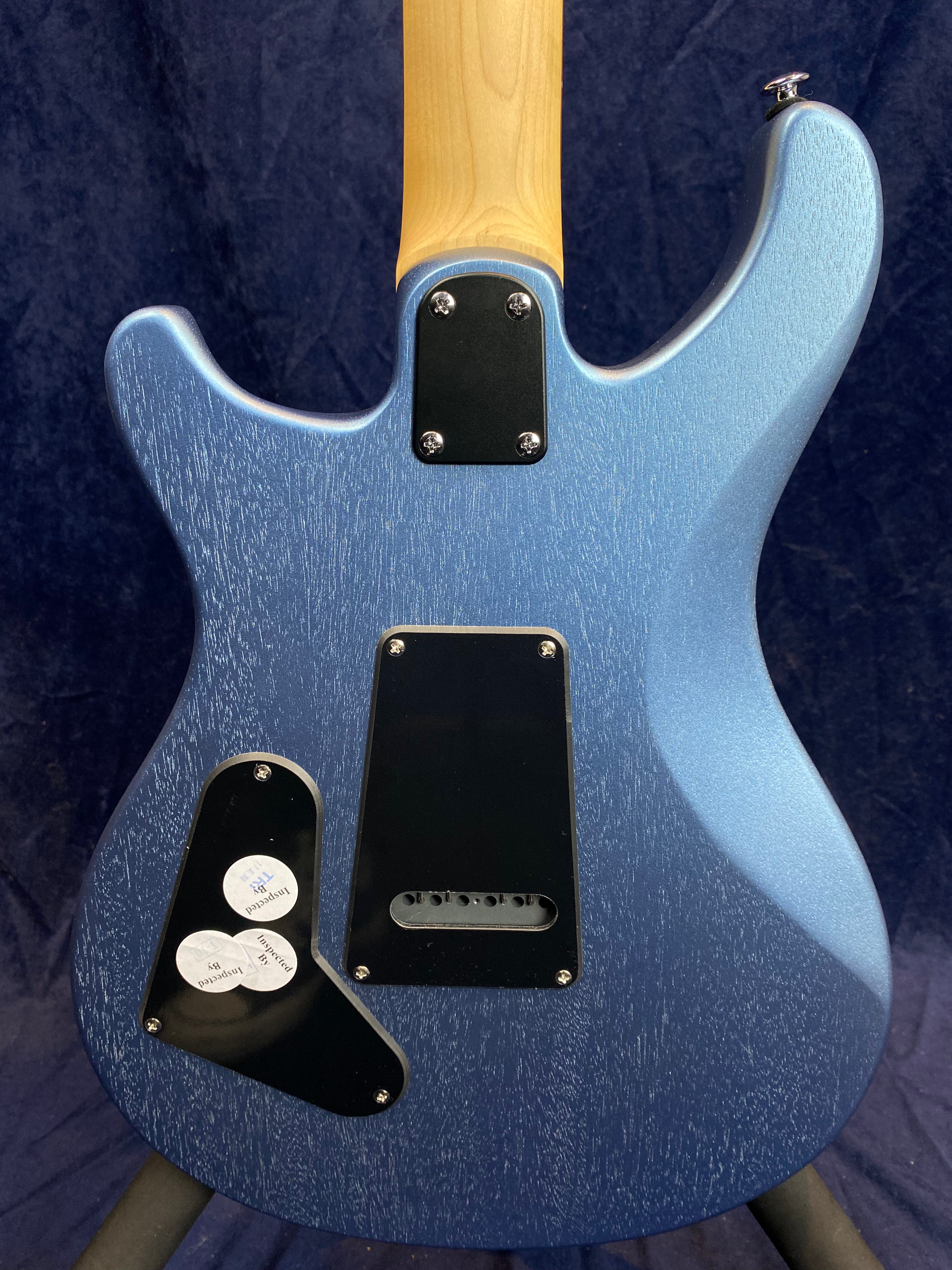 Back of a metallic blue electric guitar, showing a natural wood neck, two black access panels, and three inspection stickers on the lower panel.