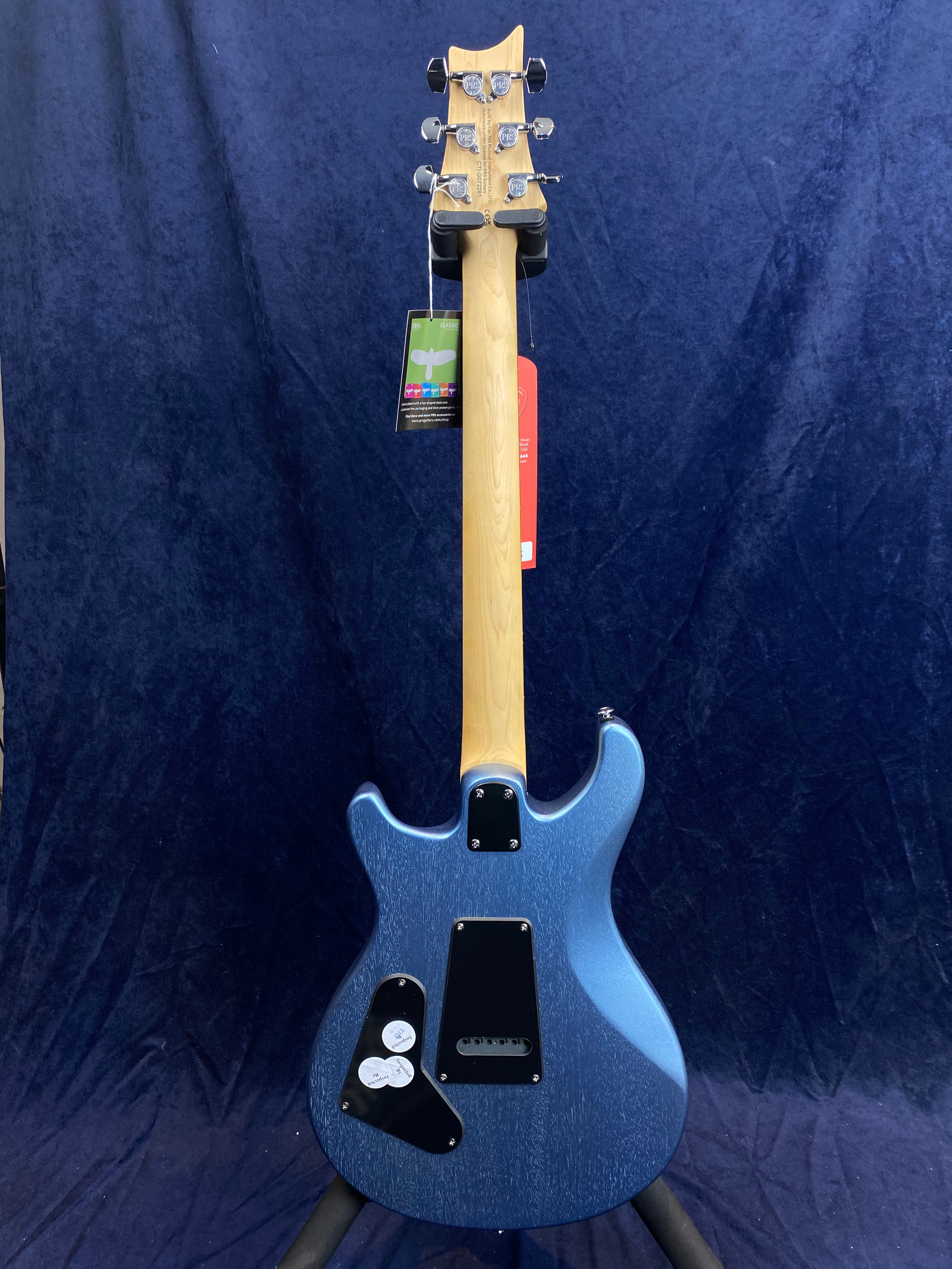 Blue electric guitar with a natural wood neck, six tuning pegs, and black plastic covers on the back of the body. Tags hang from the headstock.
