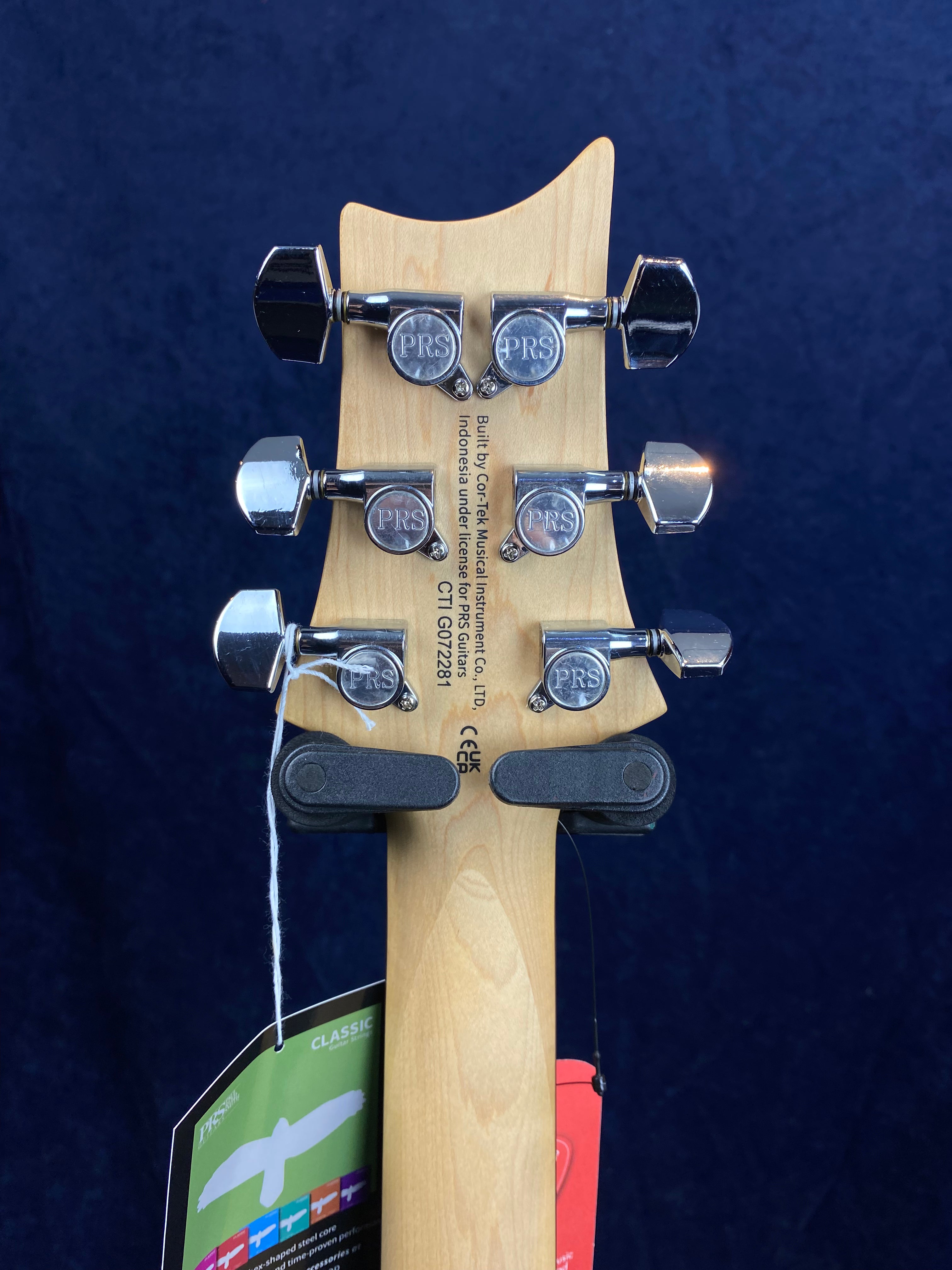Six chrome PRS-branded tuning pegs on a light wood electric guitar headstock, with two hanging tags attached below.