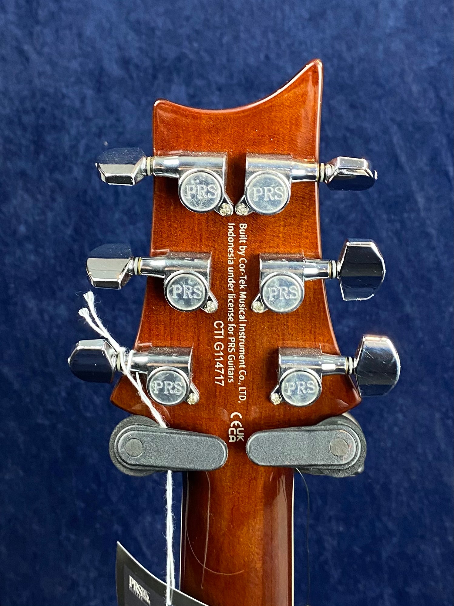 Back of a PRS electric guitar headstock showing six chrome tuning pegs, serial number, and manufacturer information on a brown wood finish.