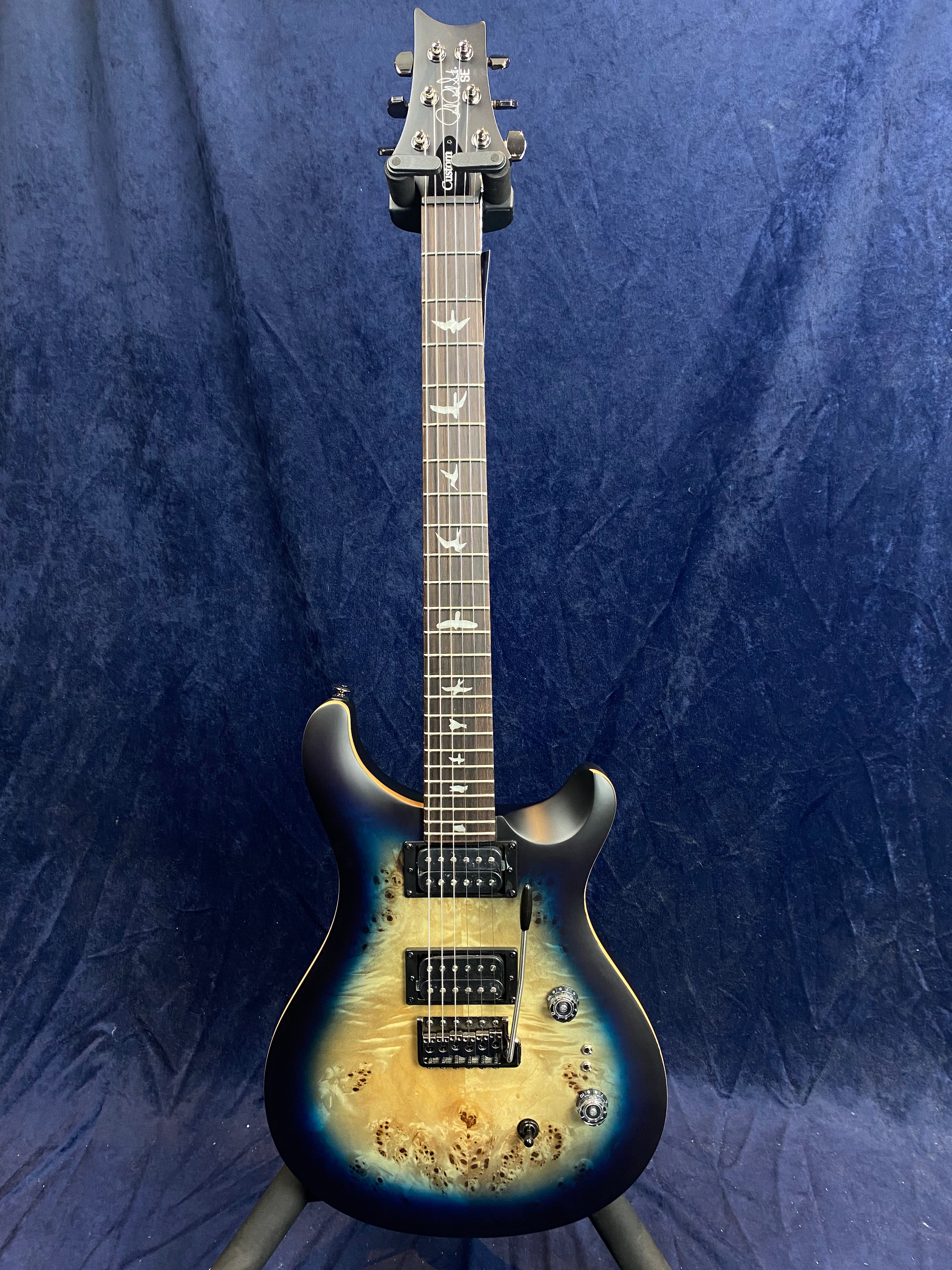 Electric guitar with a glossy blue burst finish, bird inlays on the fretboard, two pickups, and three control knobs against a dark blue background.
