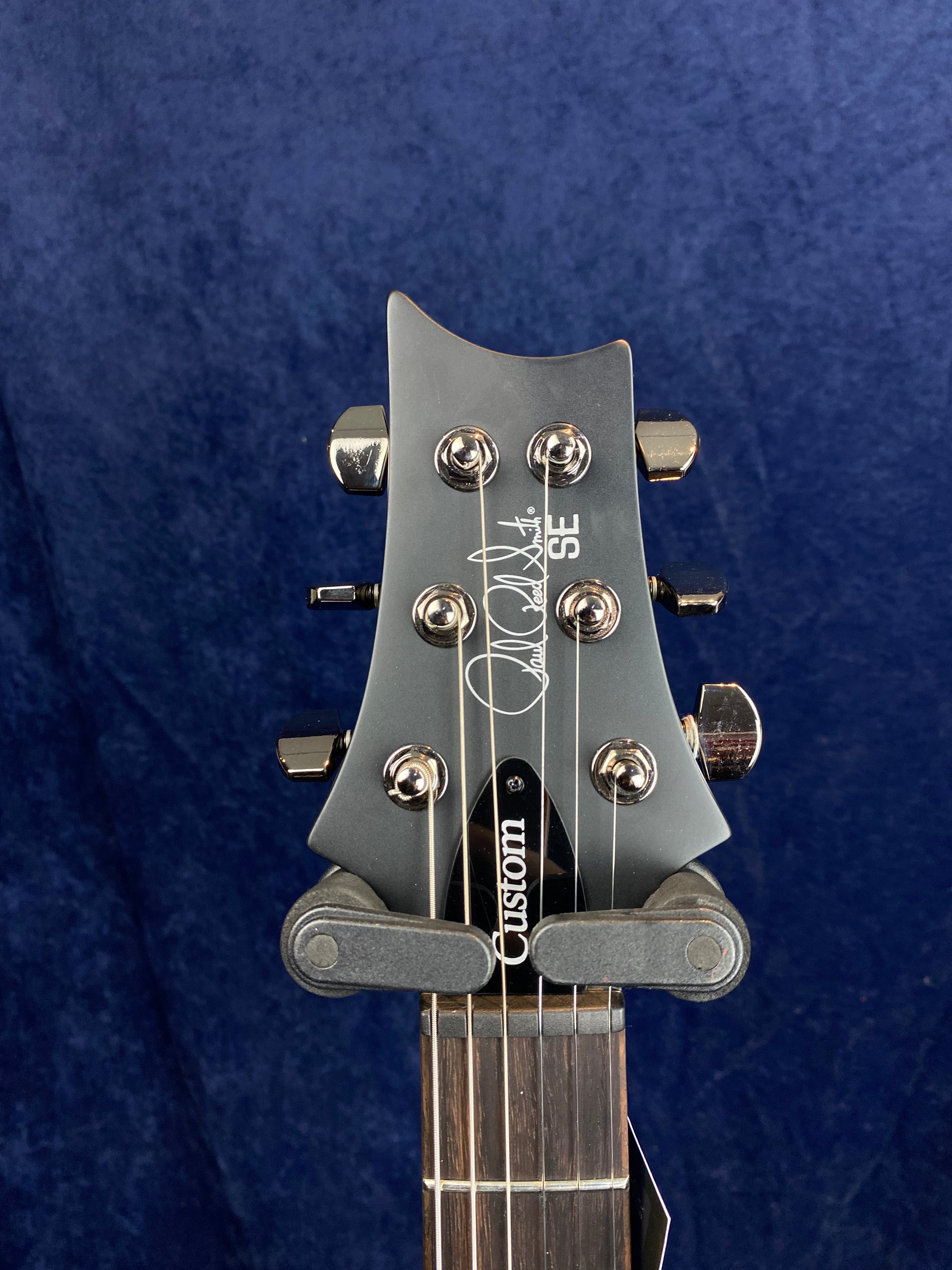 Headstock of a PRS SE Custom electric guitar with six chrome tuning pegs, black finish, and signature logo, against a dark blue background.