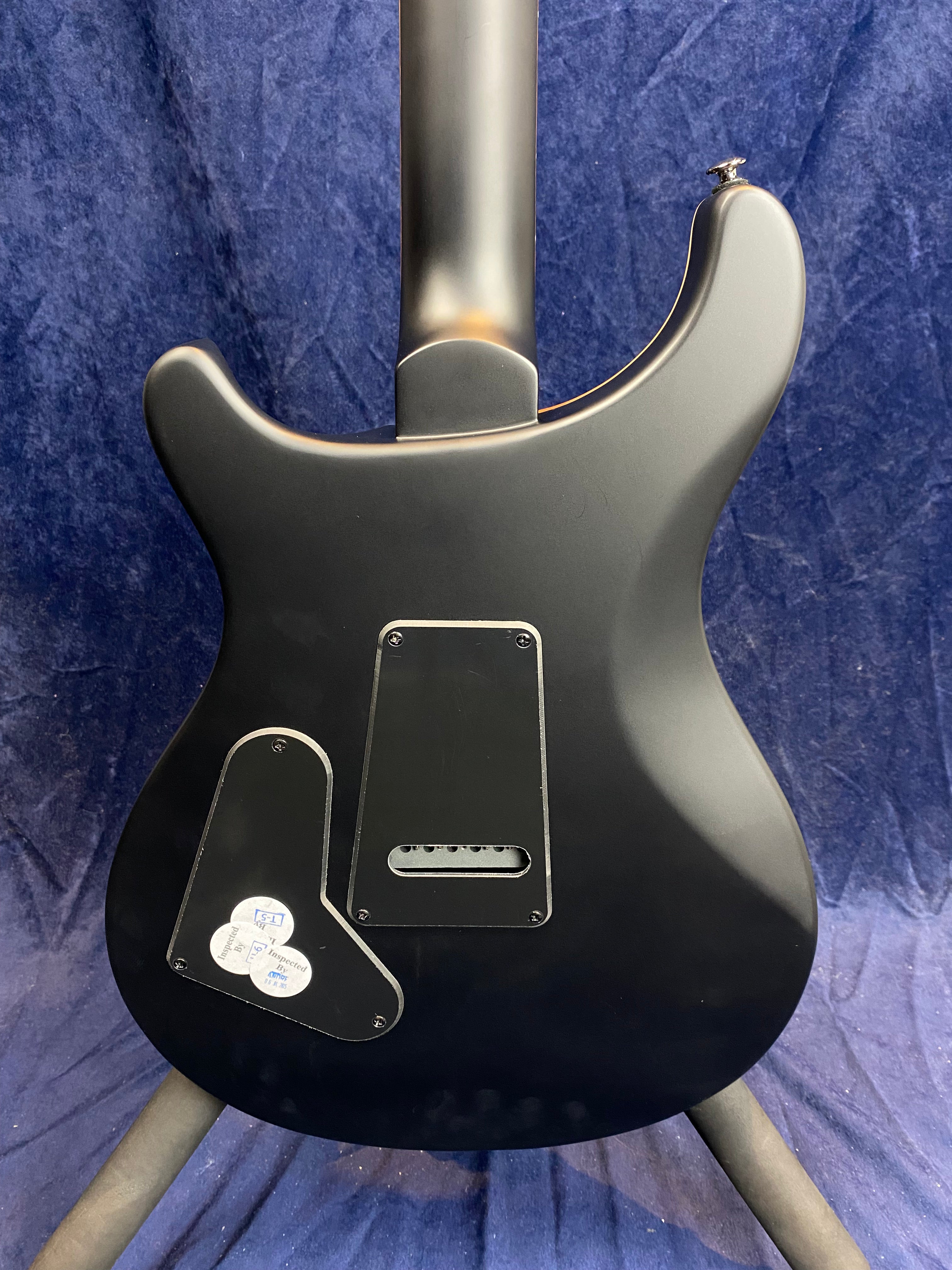 Matte black electric guitar back with two black plastic covers, one rectangular and one irregularly shaped, displayed against dark blue fabric.