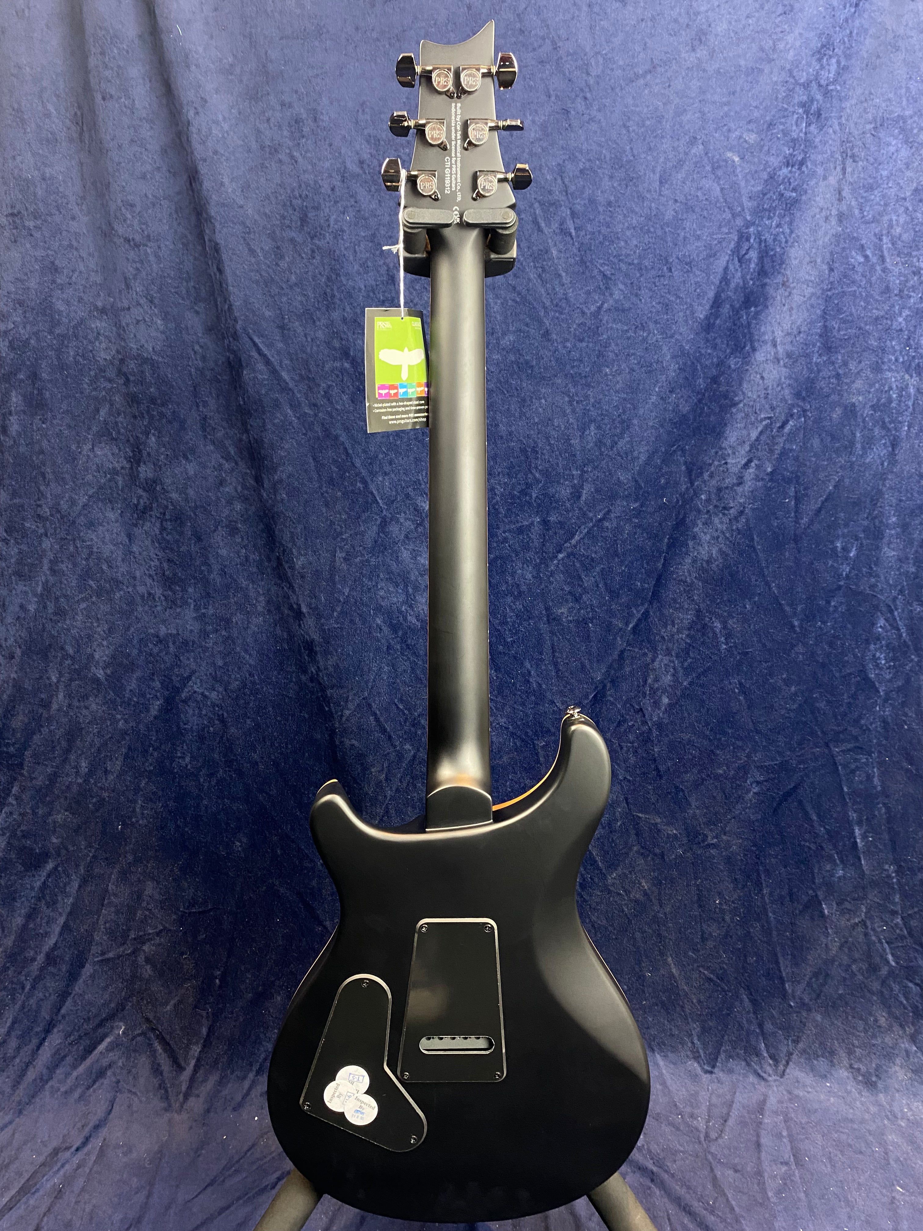 Matte black electric guitar shown from the back, with two plastic covers and a tag hanging from one tuning peg.