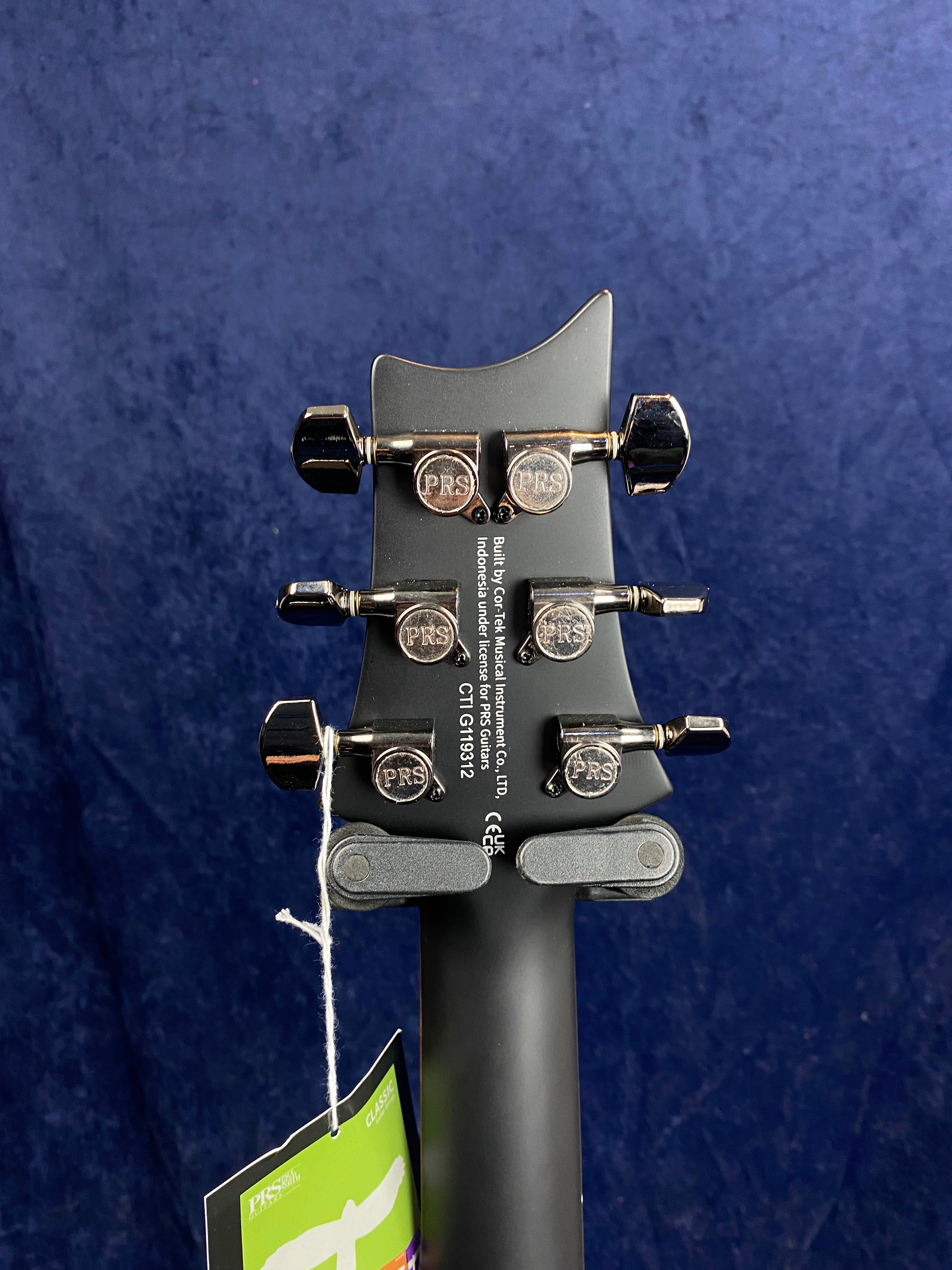 Back of a PRS electric guitar headstock with six chrome tuning pegs, PRS logo, serial number, and a tag attached by string.