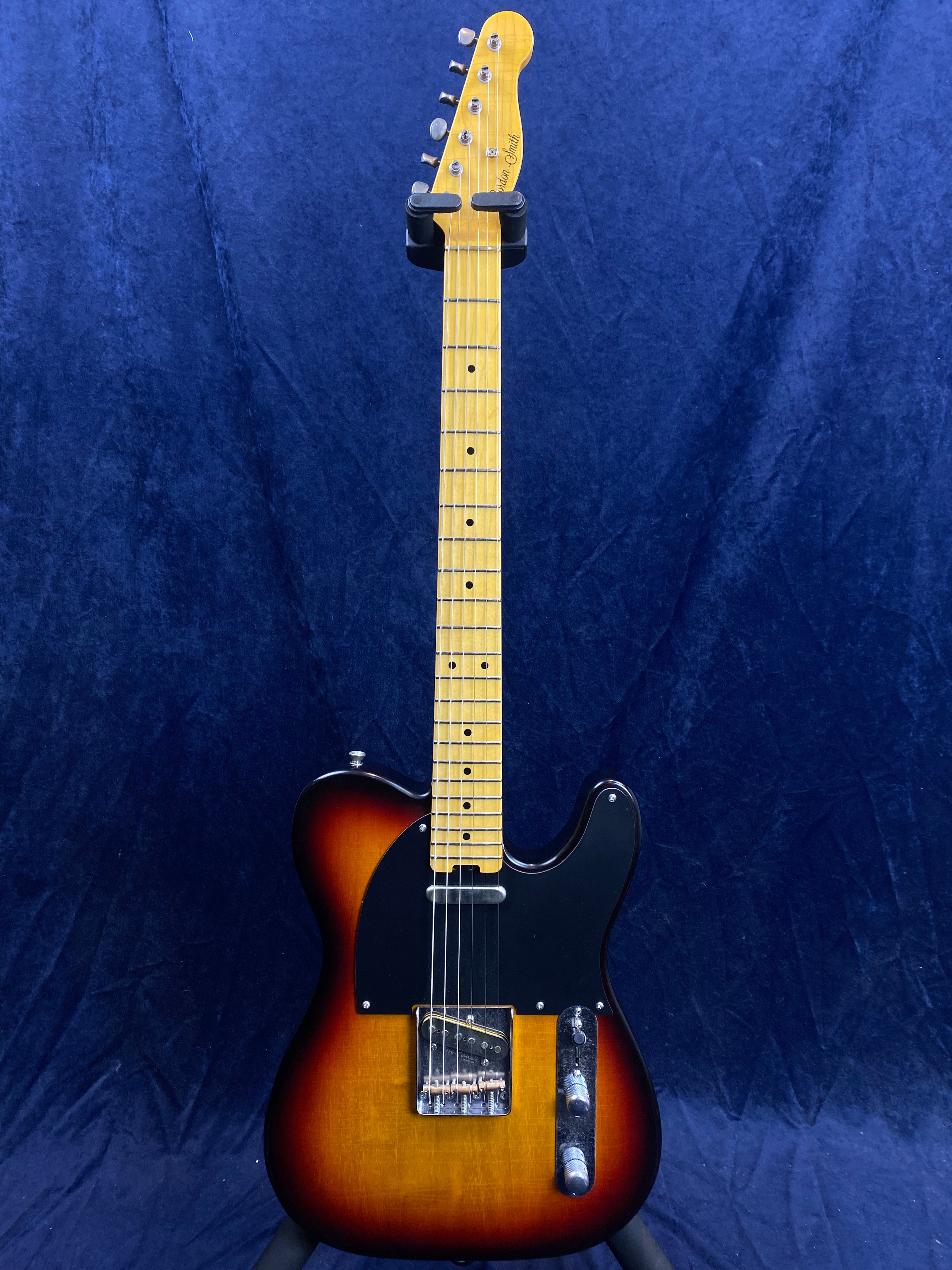 Sunburst electric guitar with a black pickguard, two chrome control knobs, single cutaway body, and a light maple neck against a dark blue background.