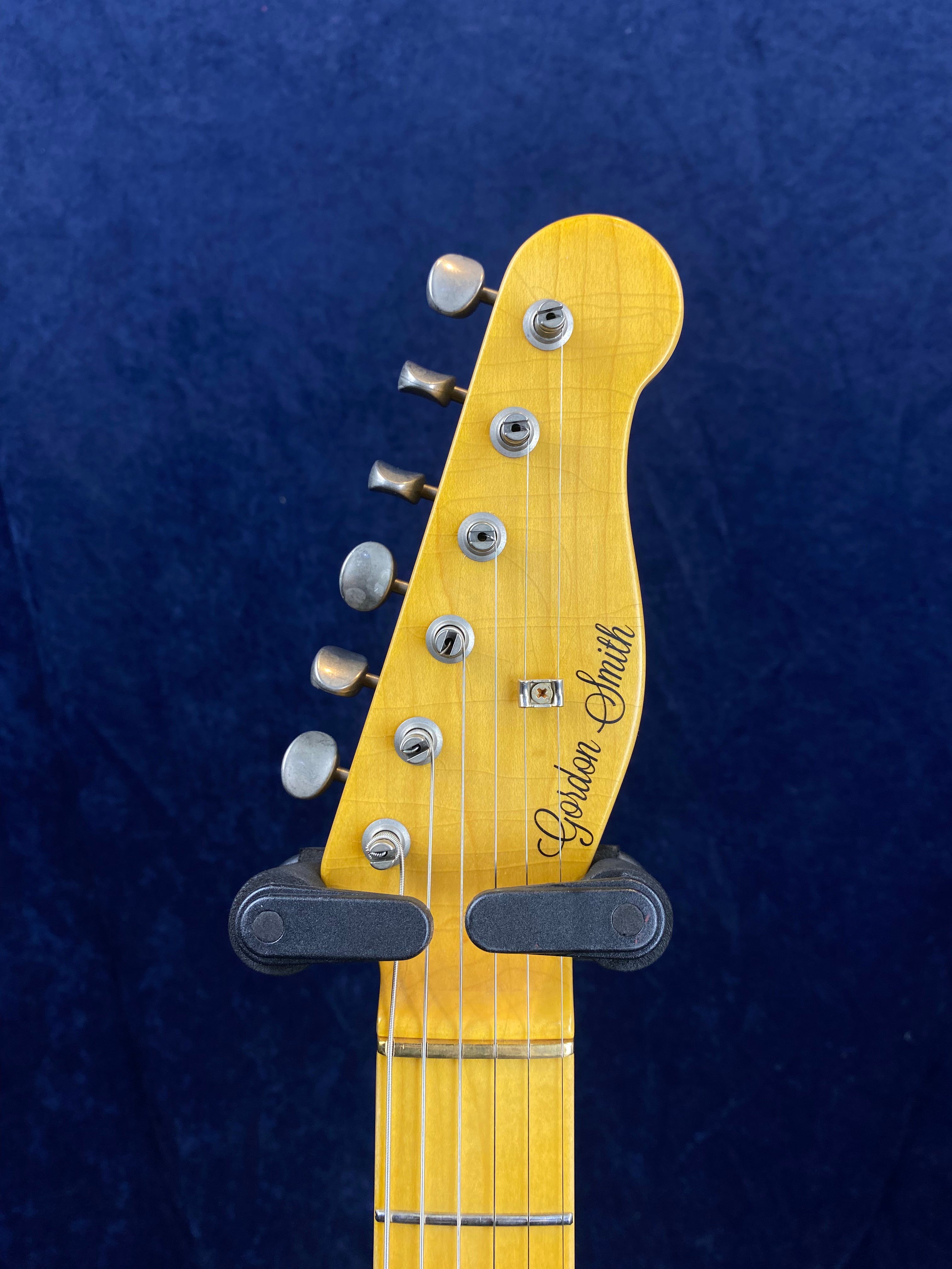 Six tuning pegs and the name Gordon Smith appear on the light wood headstock of an electric guitar, against a dark blue background.