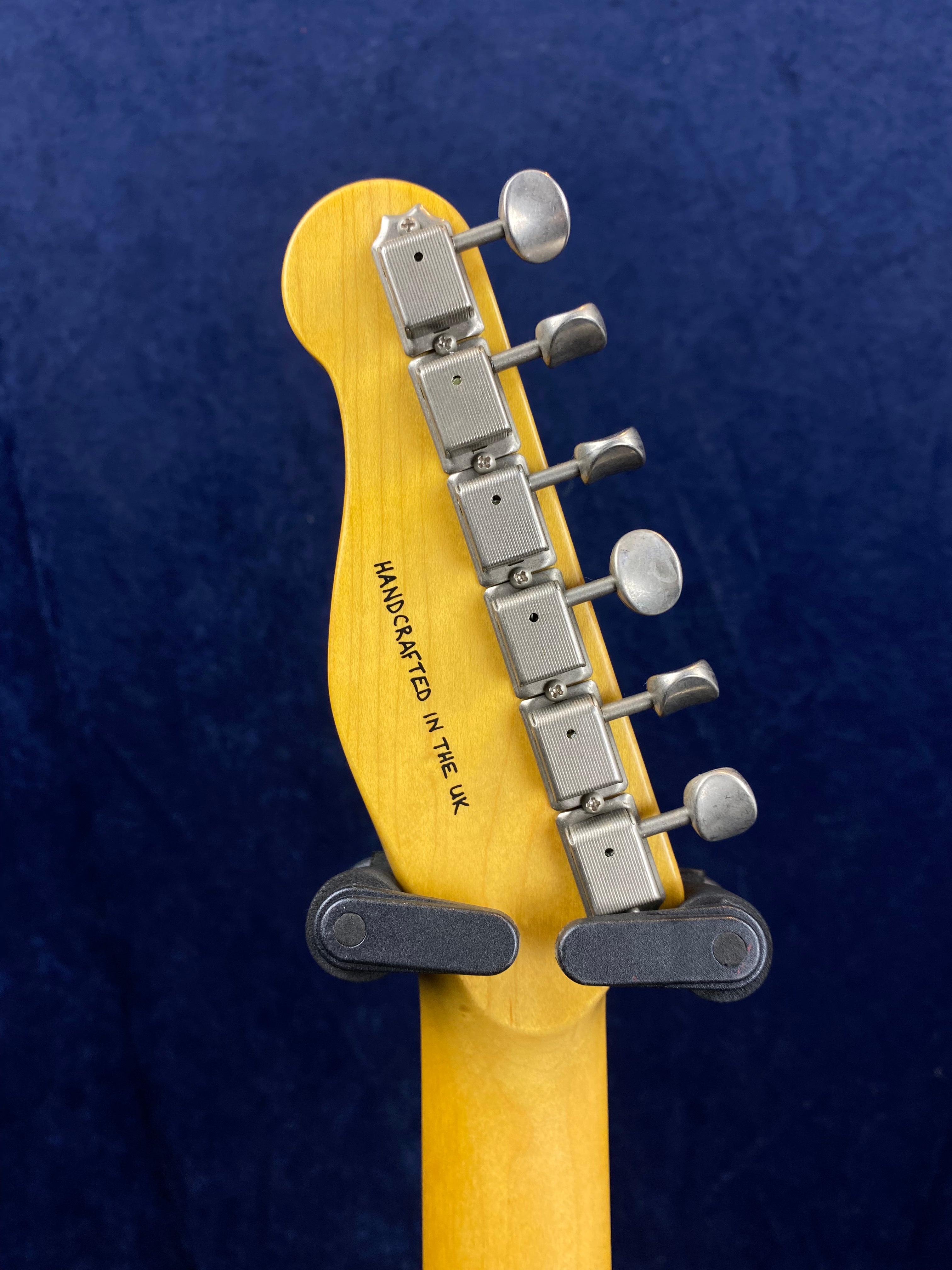Six vintage-style metal tuning pegs on a light wood electric guitar headstock, labeled "Handcrafted in the UK" against a dark blue background.