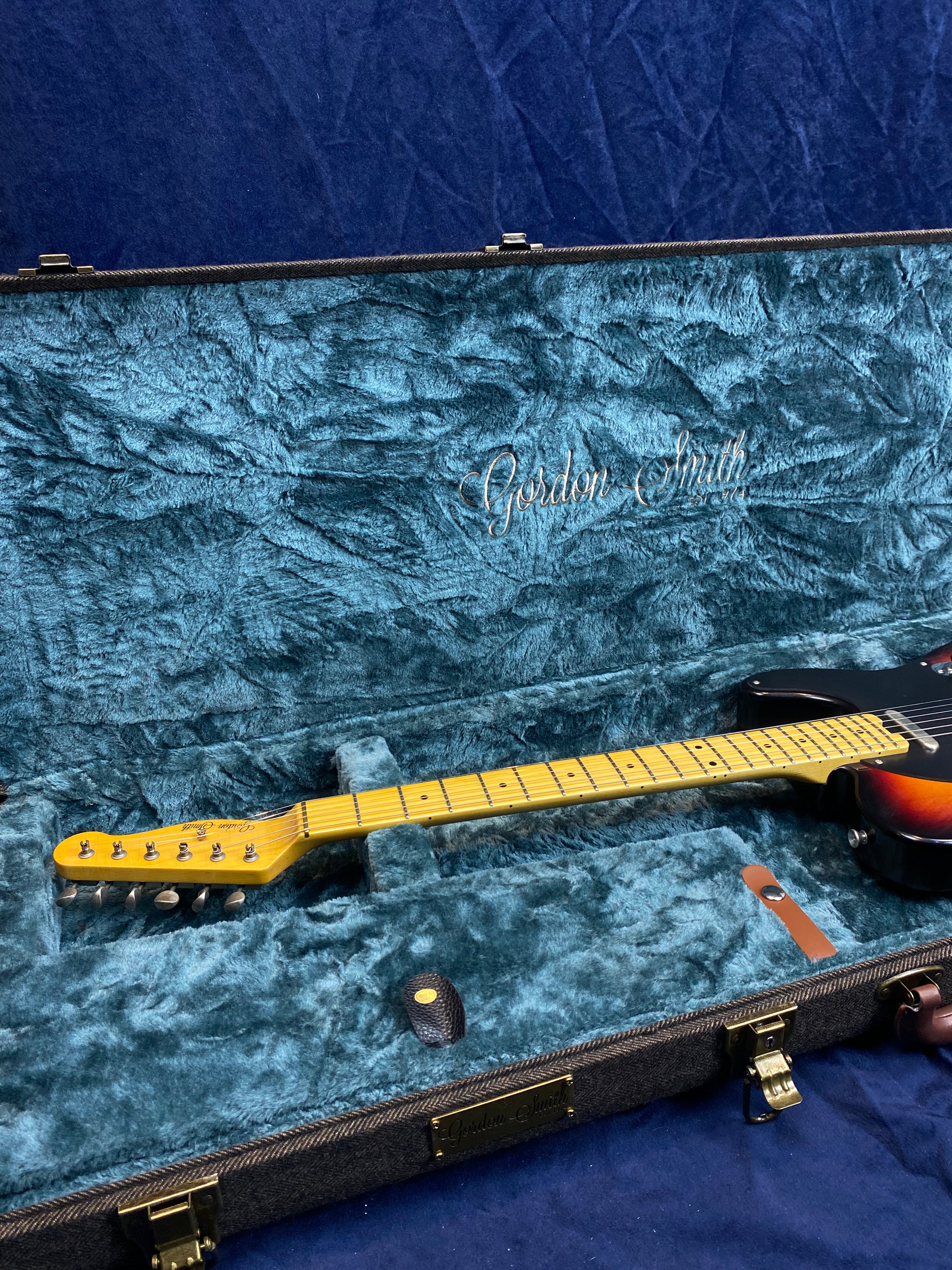 Electric guitar with a sunburst finish and maple neck resting in a blue velvet-lined Gordon Smith hard case.