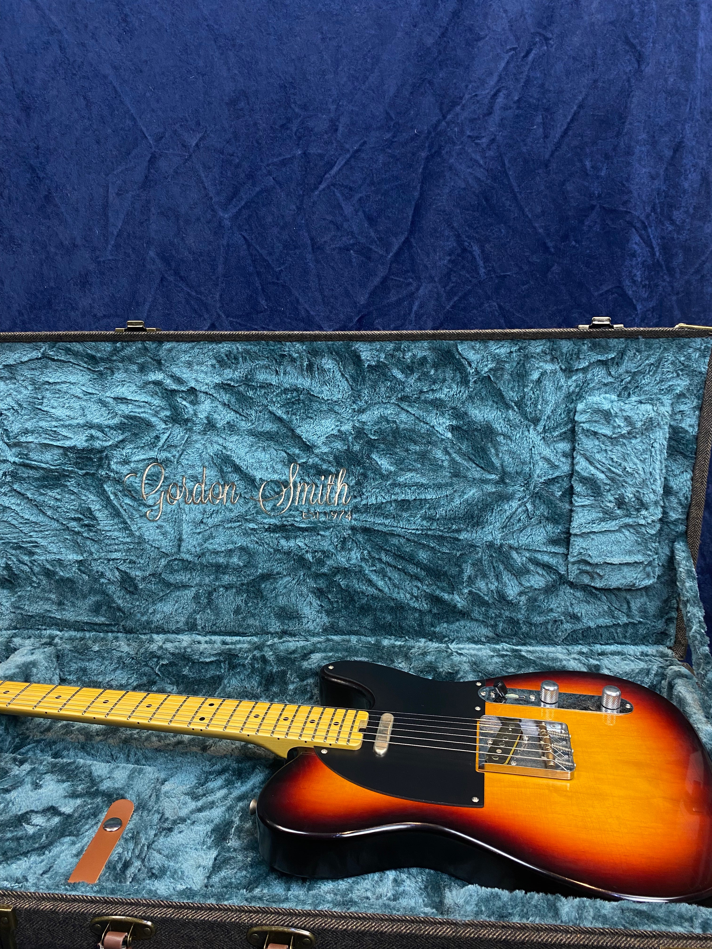 Sunburst-finish electric guitar with black pickguard in a teal-lined hard case, labeled "Gordon Smith" inside the lid.