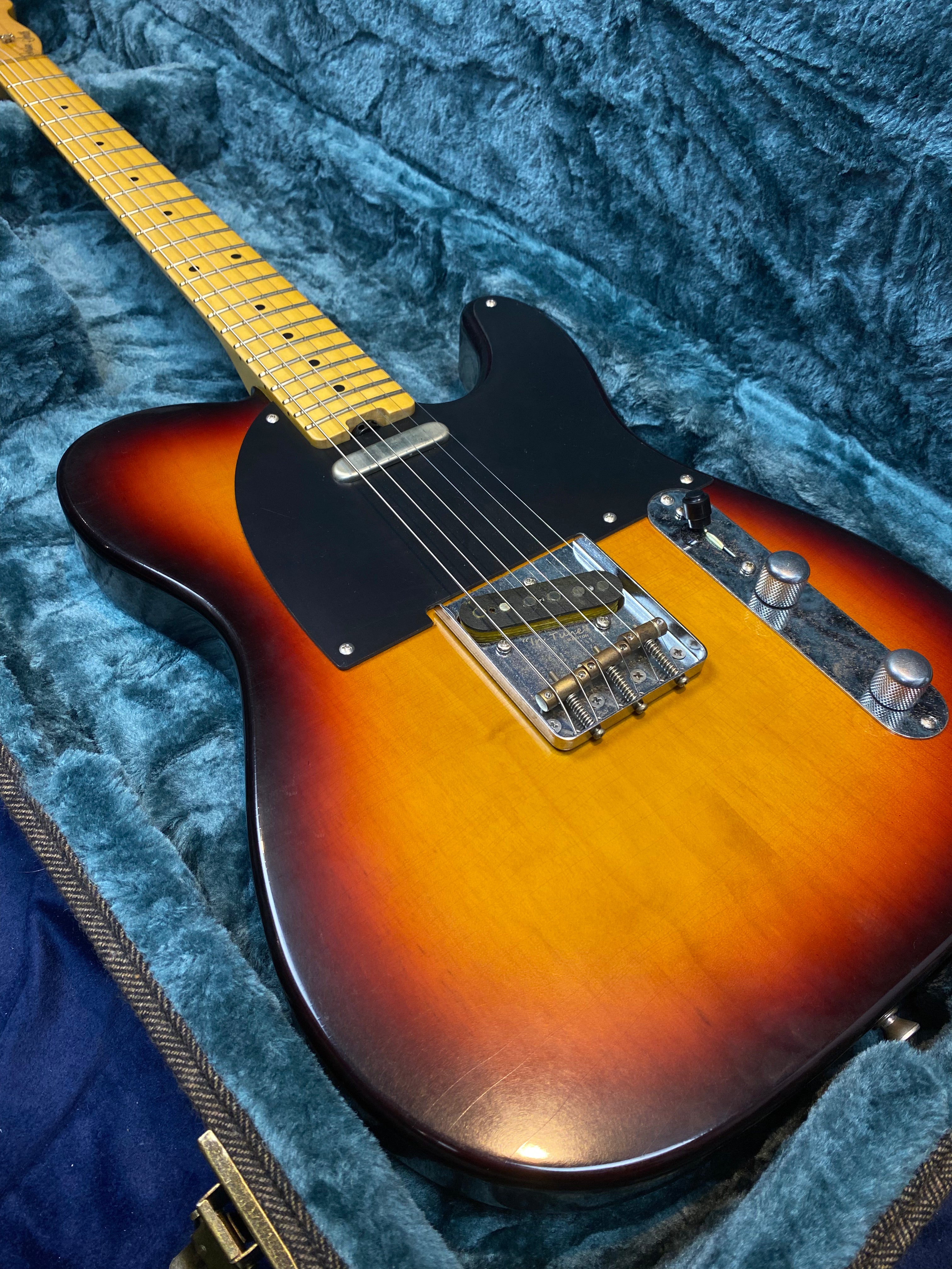 Sunburst electric guitar with a black pickguard, chrome bridge, two control knobs, and a maple neck resting in a plush-lined guitar case.