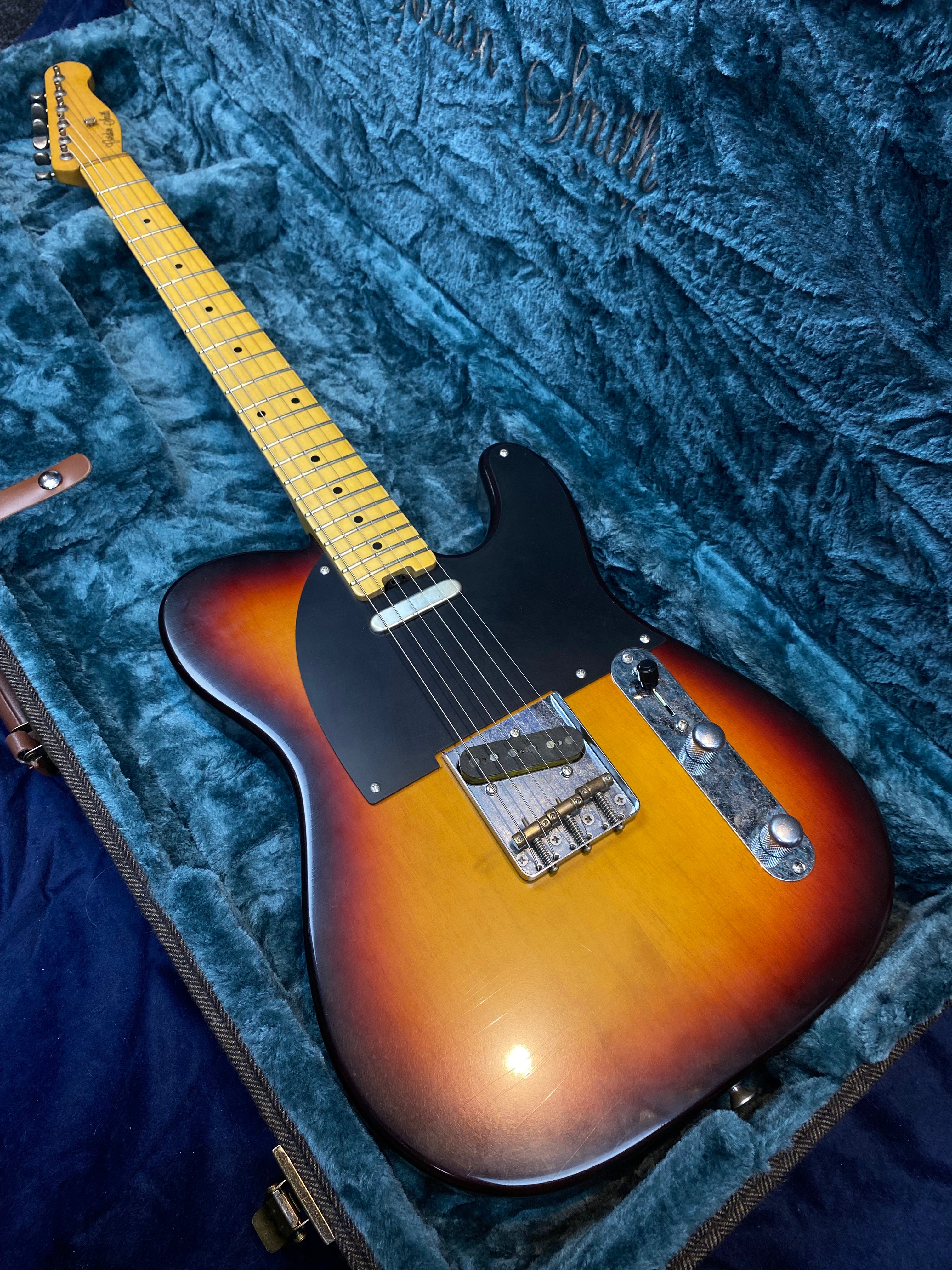 Sunburst electric guitar with a black pickguard, maple neck, and two control knobs, resting inside a blue plush-lined hard case.