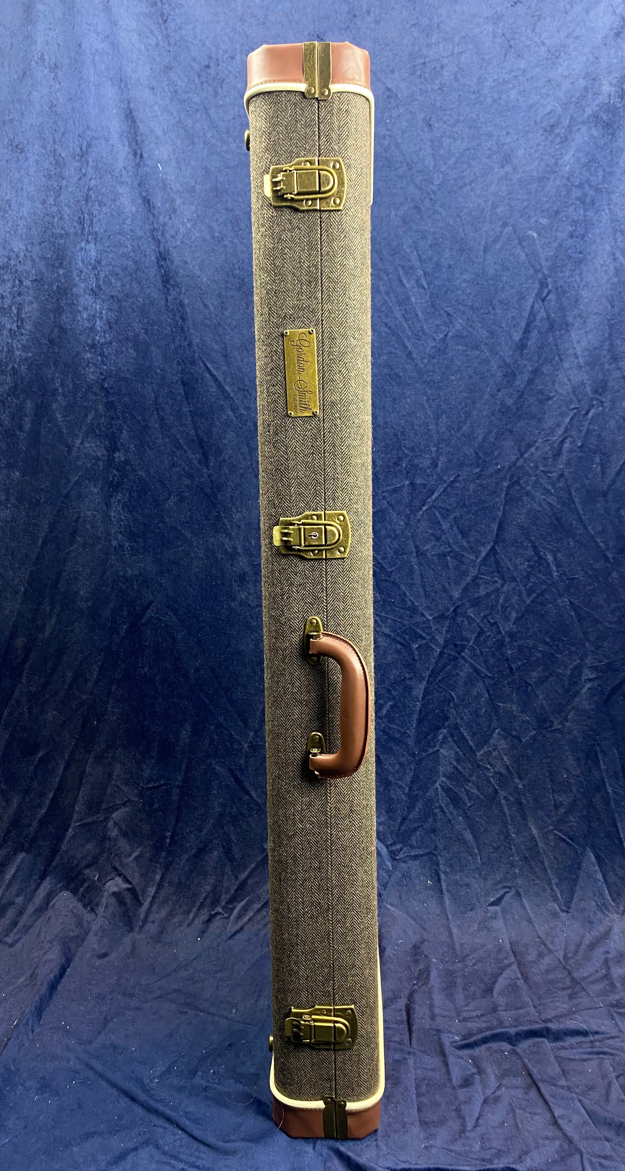 Tall, rectangular tweed electric guitar case with brown leather corners, brass latches, a brown handle, and a metal logo plate on the side.