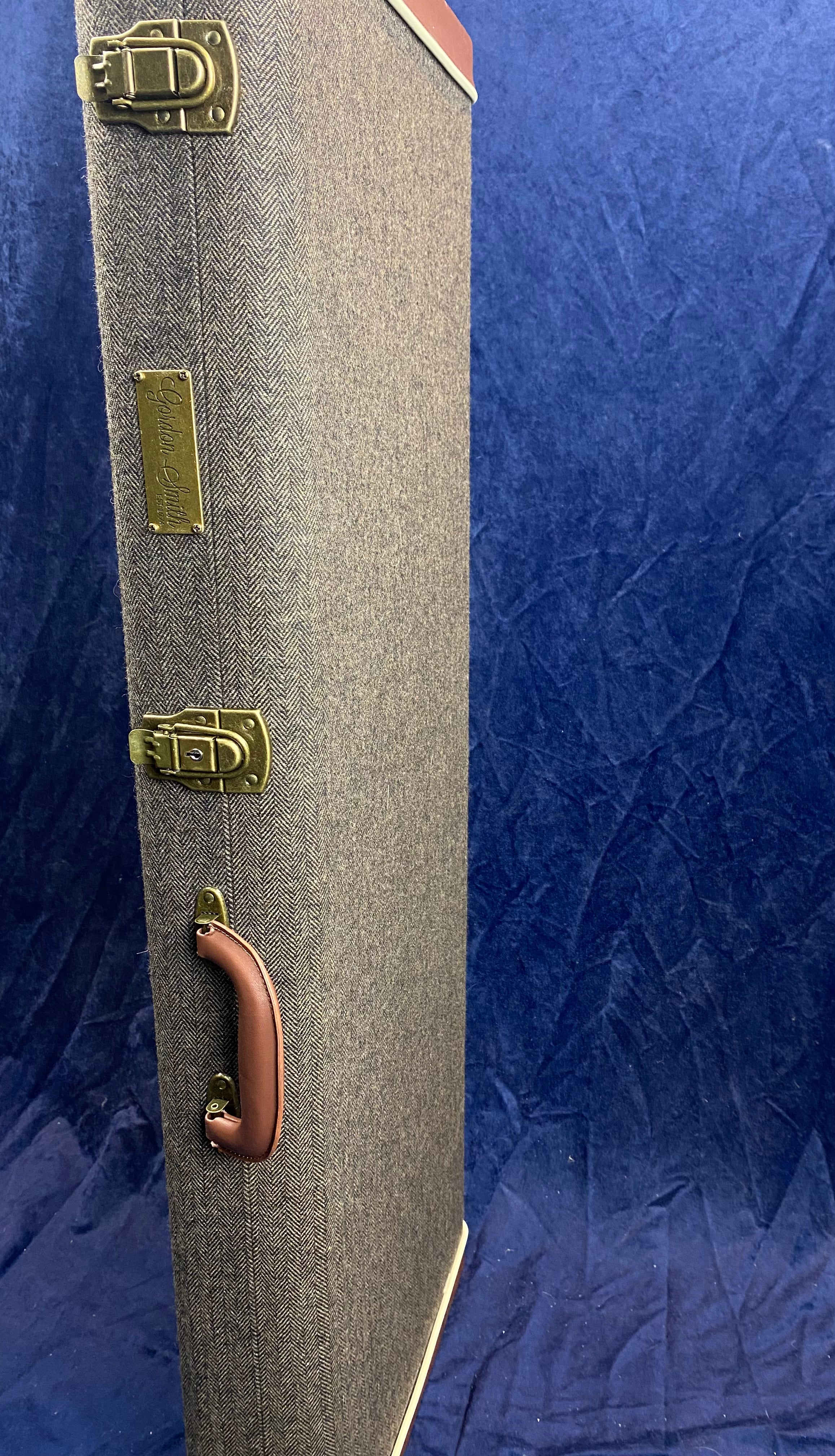 Tall rectangular electric guitar case with gray herringbone fabric, brass latches, brown leather handle, and a metal Gibson label against a blue backdrop.