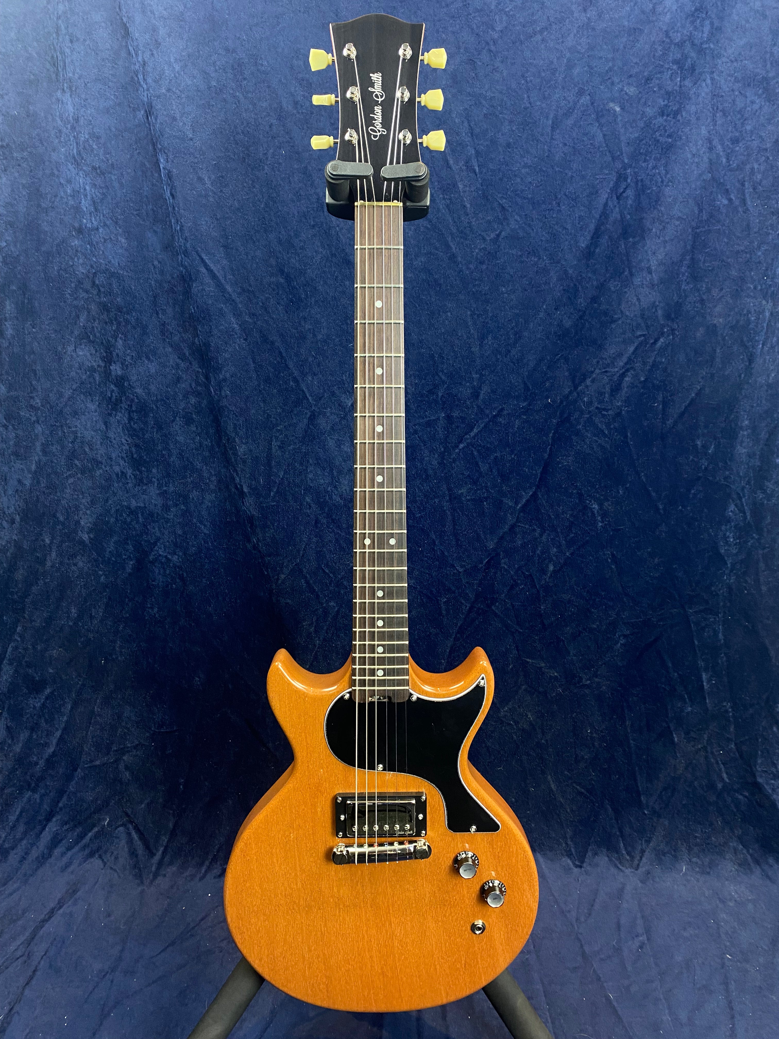 Single-cutaway electric guitar with a natural wood finish, black pickguard, one pickup, two control knobs, and yellow tuning pegs.