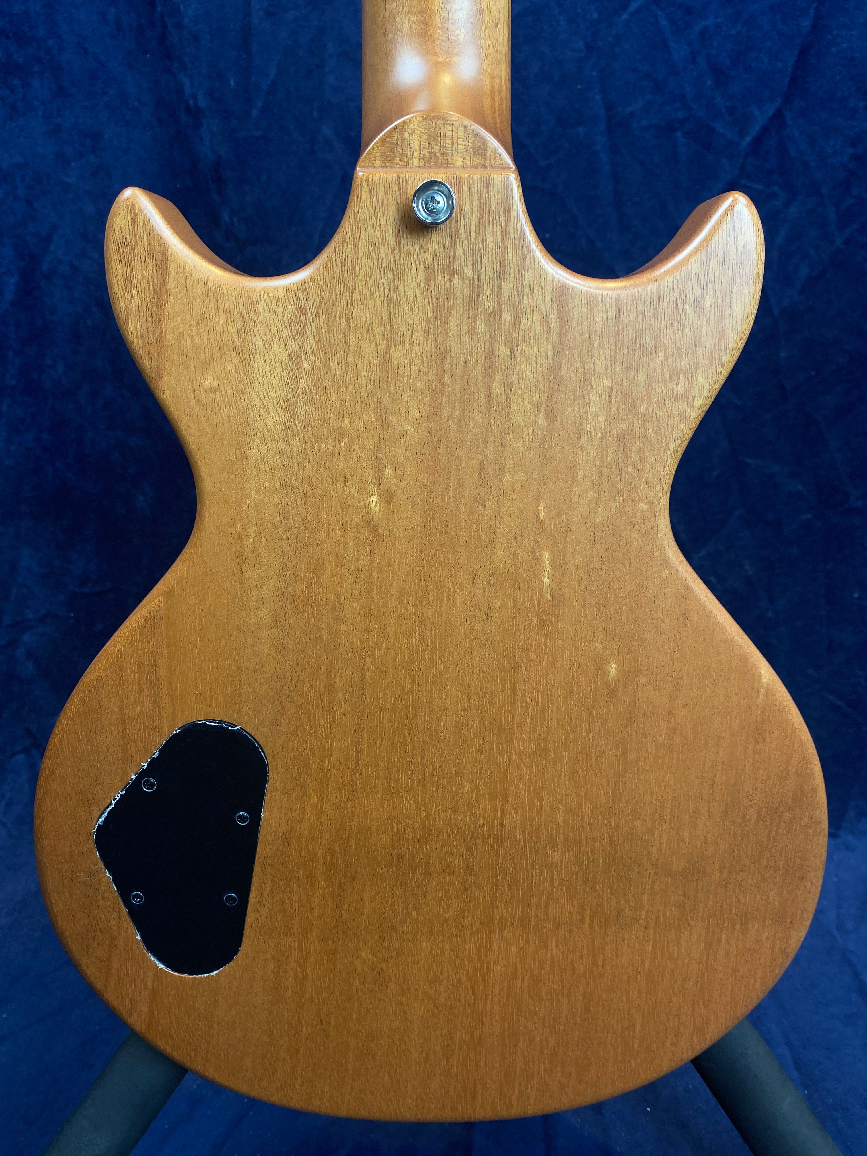 Back of an electric guitar with natural wood finish, black electronics cover plate on lower left, and a strap button near the neck joint.