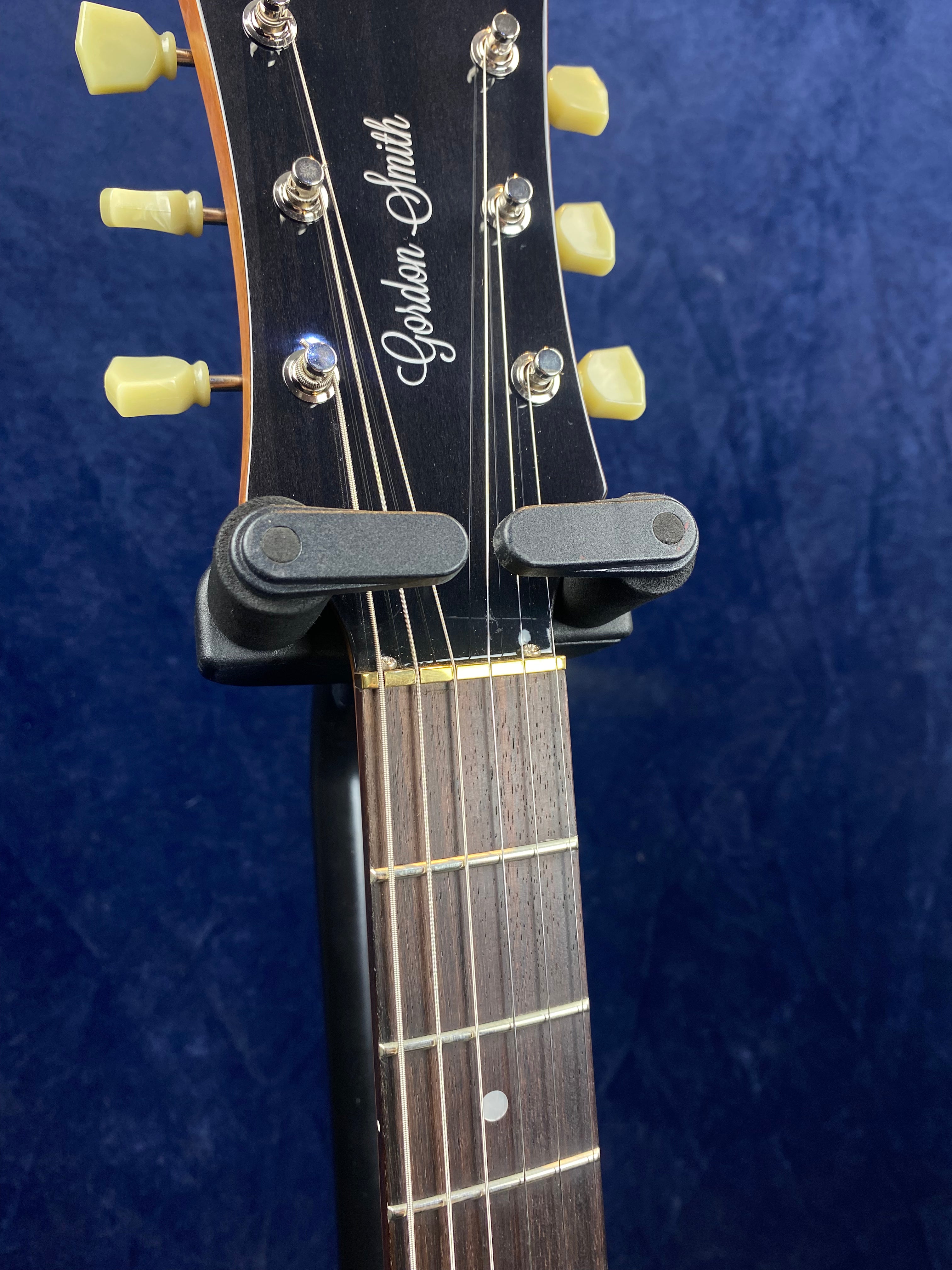Gordon Smith electric guitar headstock with cream tuning pegs and part of the fretboard, resting in a black guitar hanger against a dark blue background.