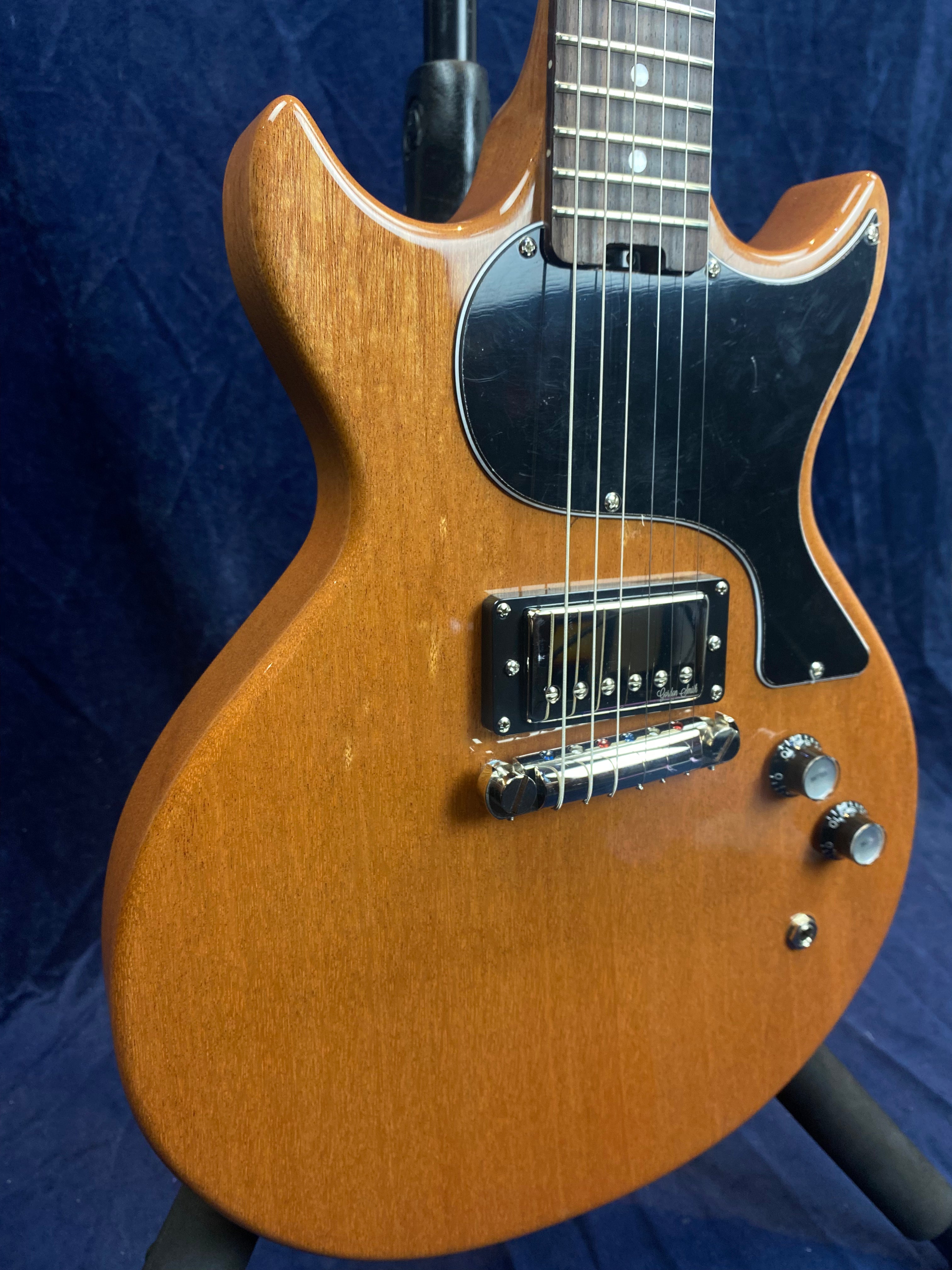 Natural wood finish electric guitar with a double cutaway body, black pickguard, single humbucker pickup, two control knobs, and metal bridge.