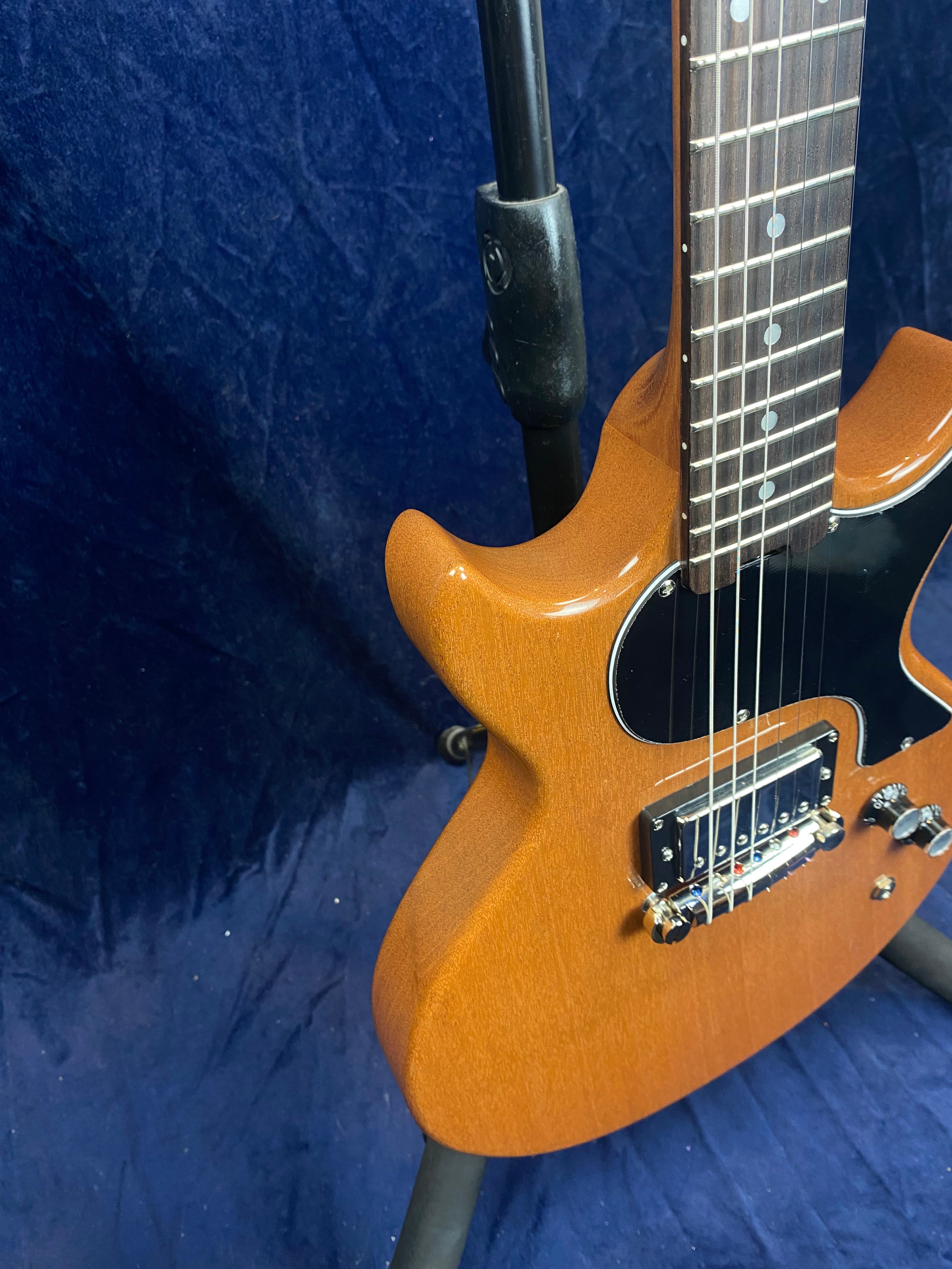 Brown electric guitar with a single black pickup, black pickguard, and silver control knobs, shown on a stand against a blue background.
