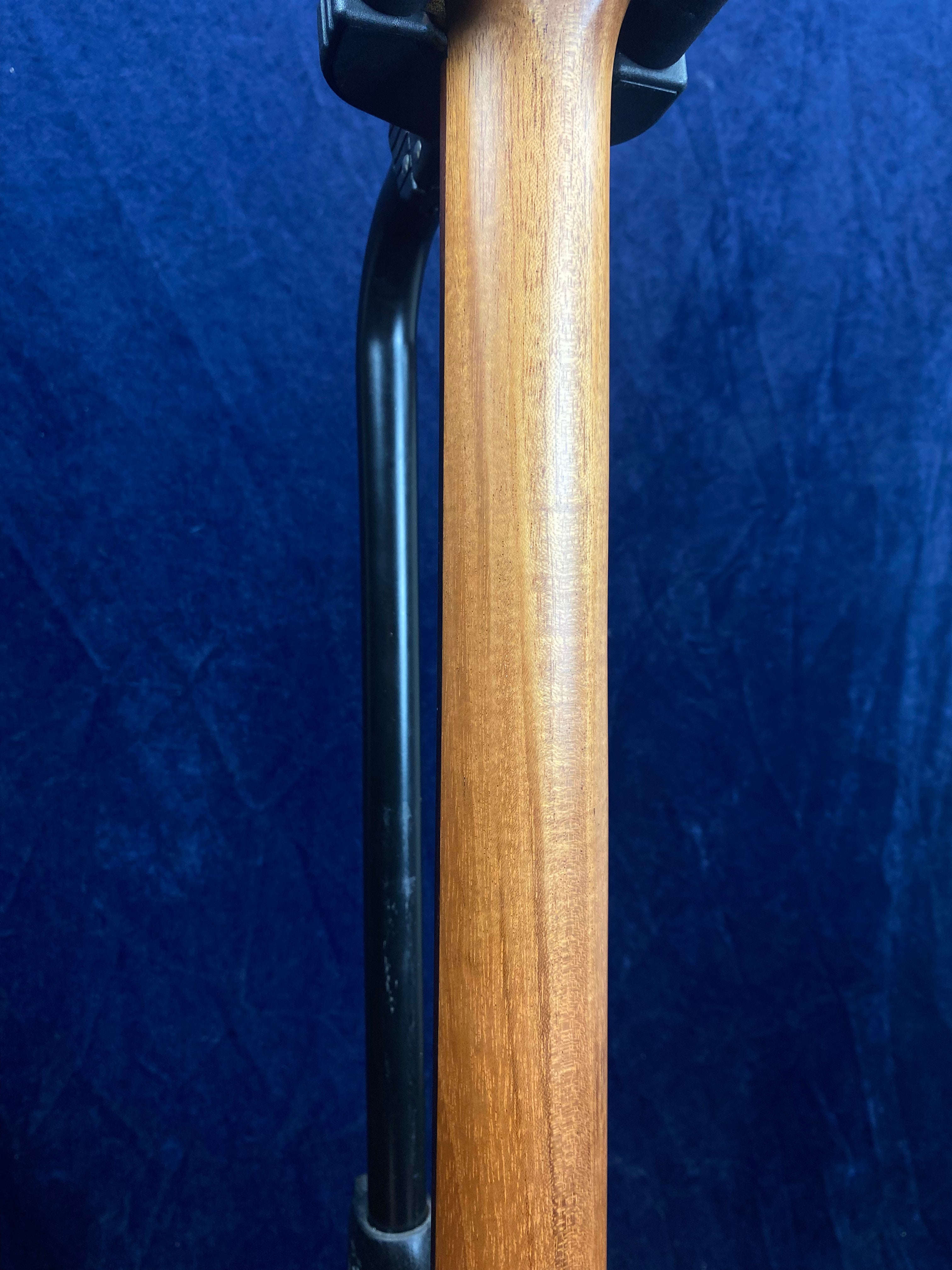 Close-up of an electric guitar neck made of smooth brown wood, mounted on a black stand against a dark blue background.