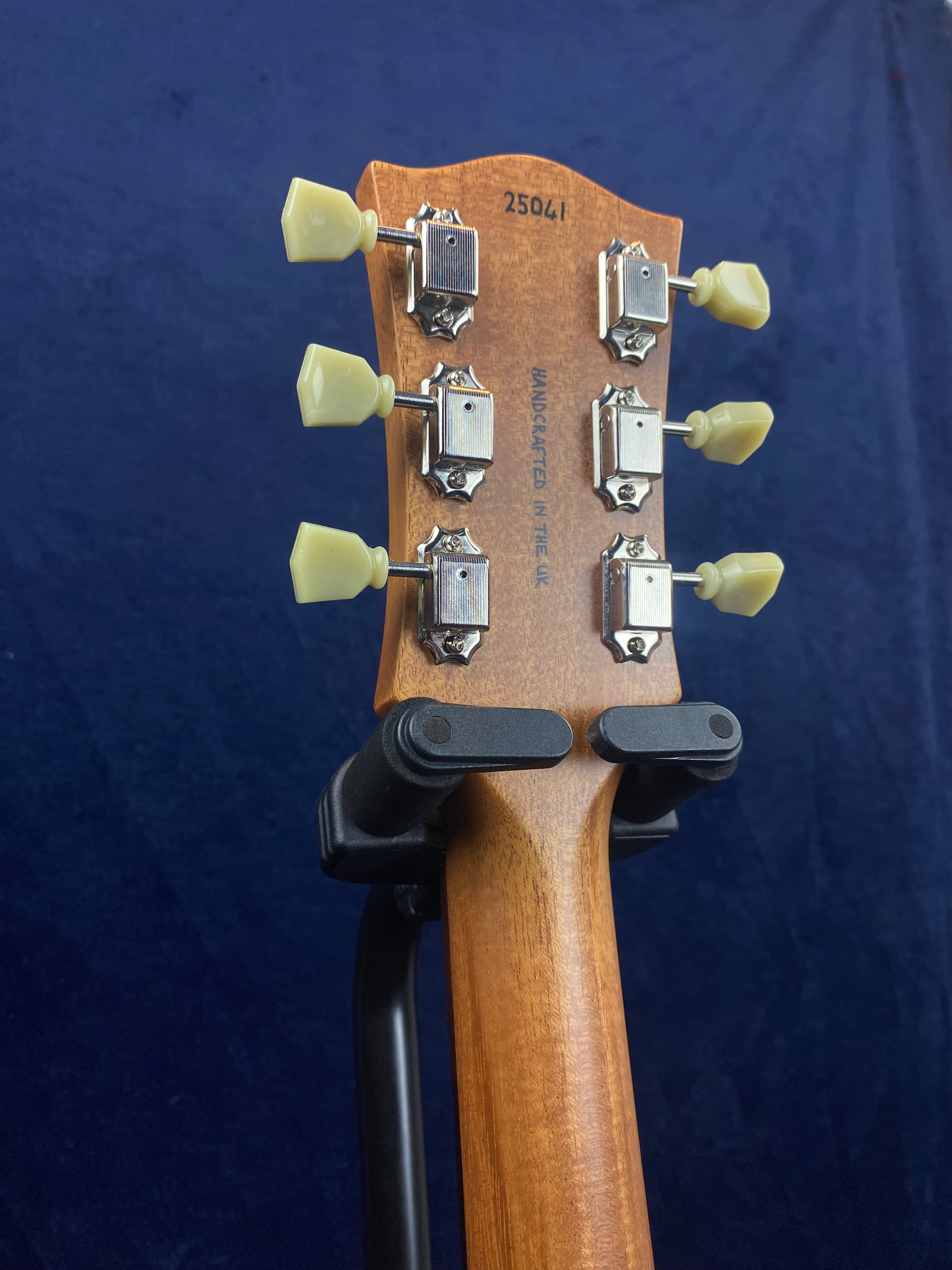 Six tuning pegs with cream knobs on a wooden electric guitar headstock, serial number 25041 and "Handcrafted in the UK" stamped on the back.